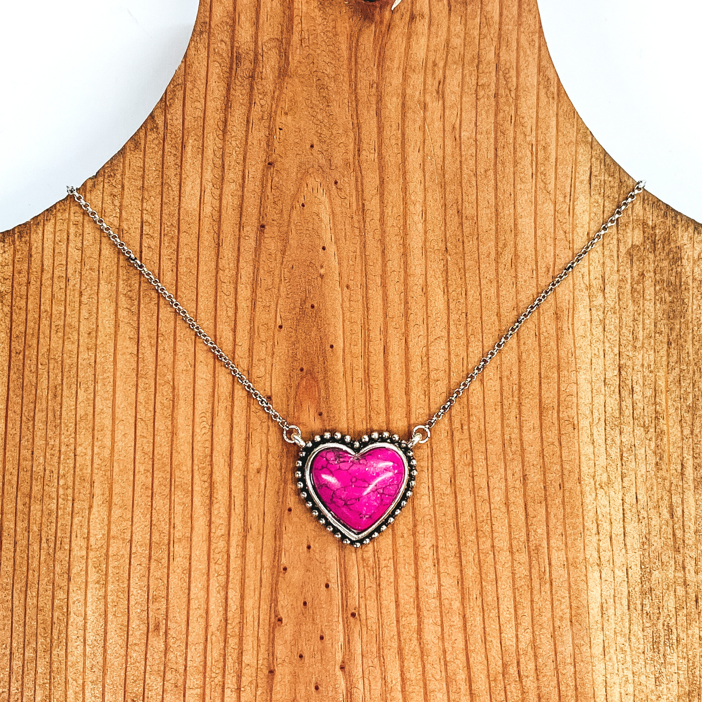 Silver chained necklace with a small pink, heart shaped stone pendant. This necklace is pictured laying on a brown necklace holder on a white and black background.