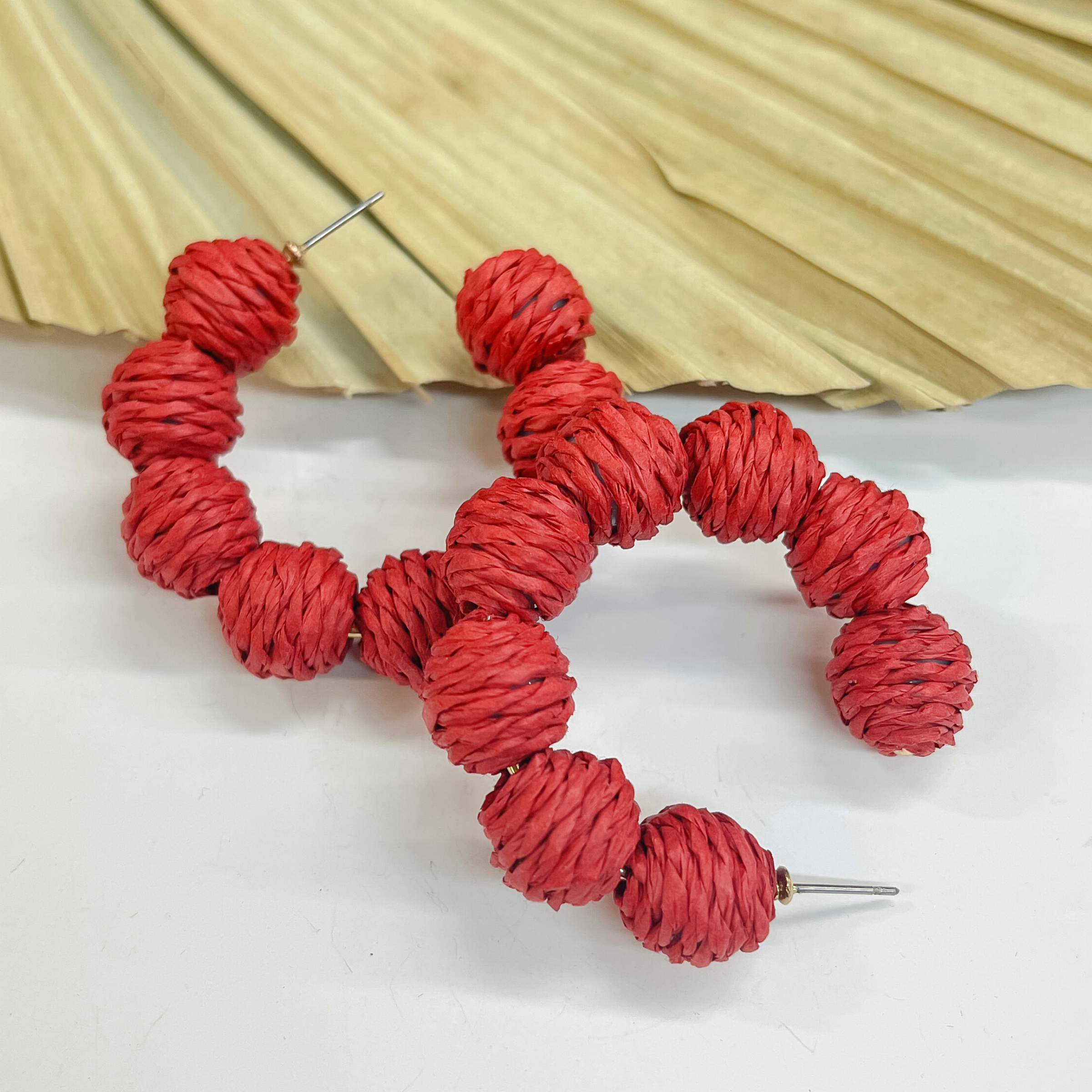 Hoop earrings with raffia balls around. The balls are wrapped in red raffia. Taken on a white background and  leaned up against a dried up palm leaf.
