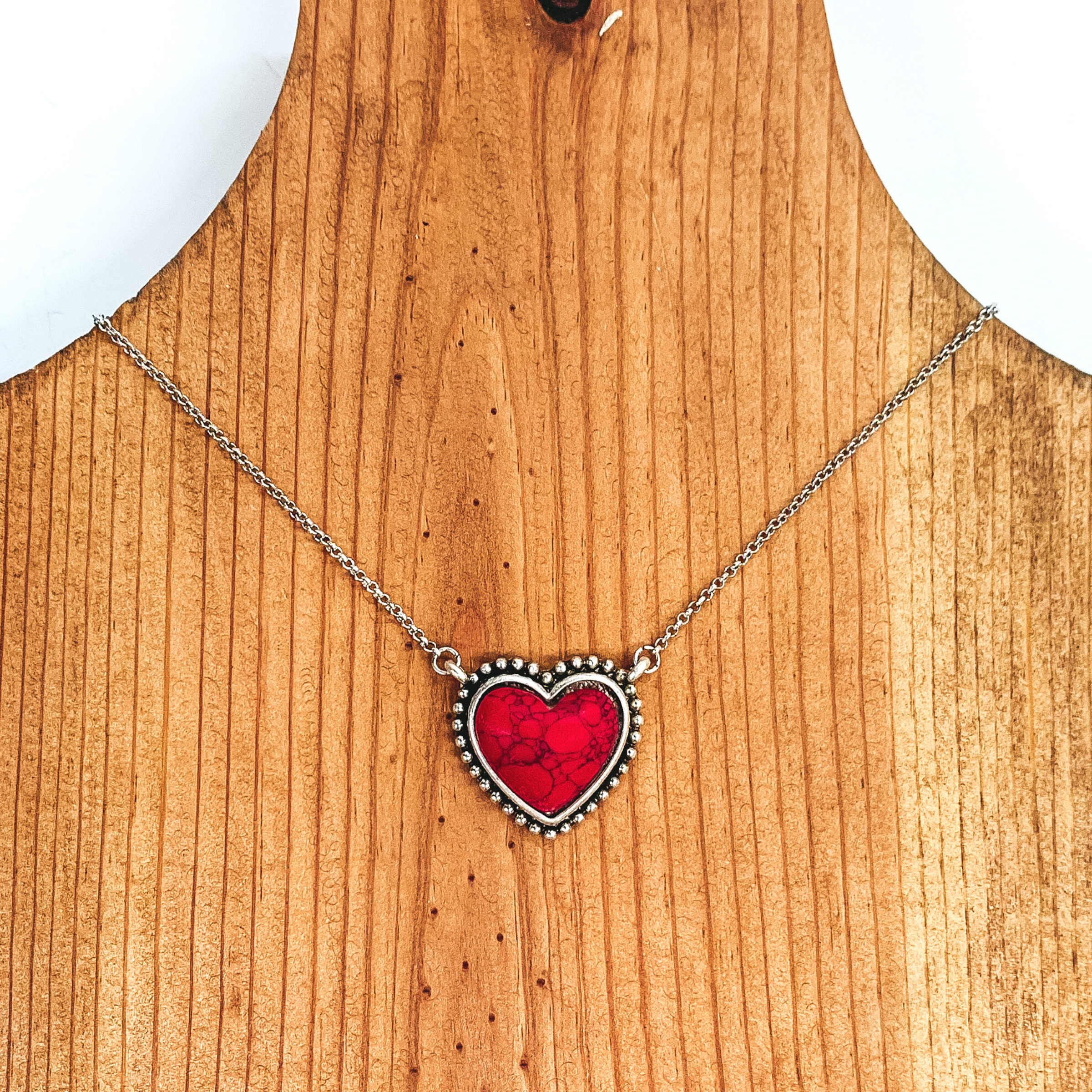 Silver chained necklace with a small red, heart shaped stone pendant. This necklace is pictured laying on a brown necklace holder on a white and black background.