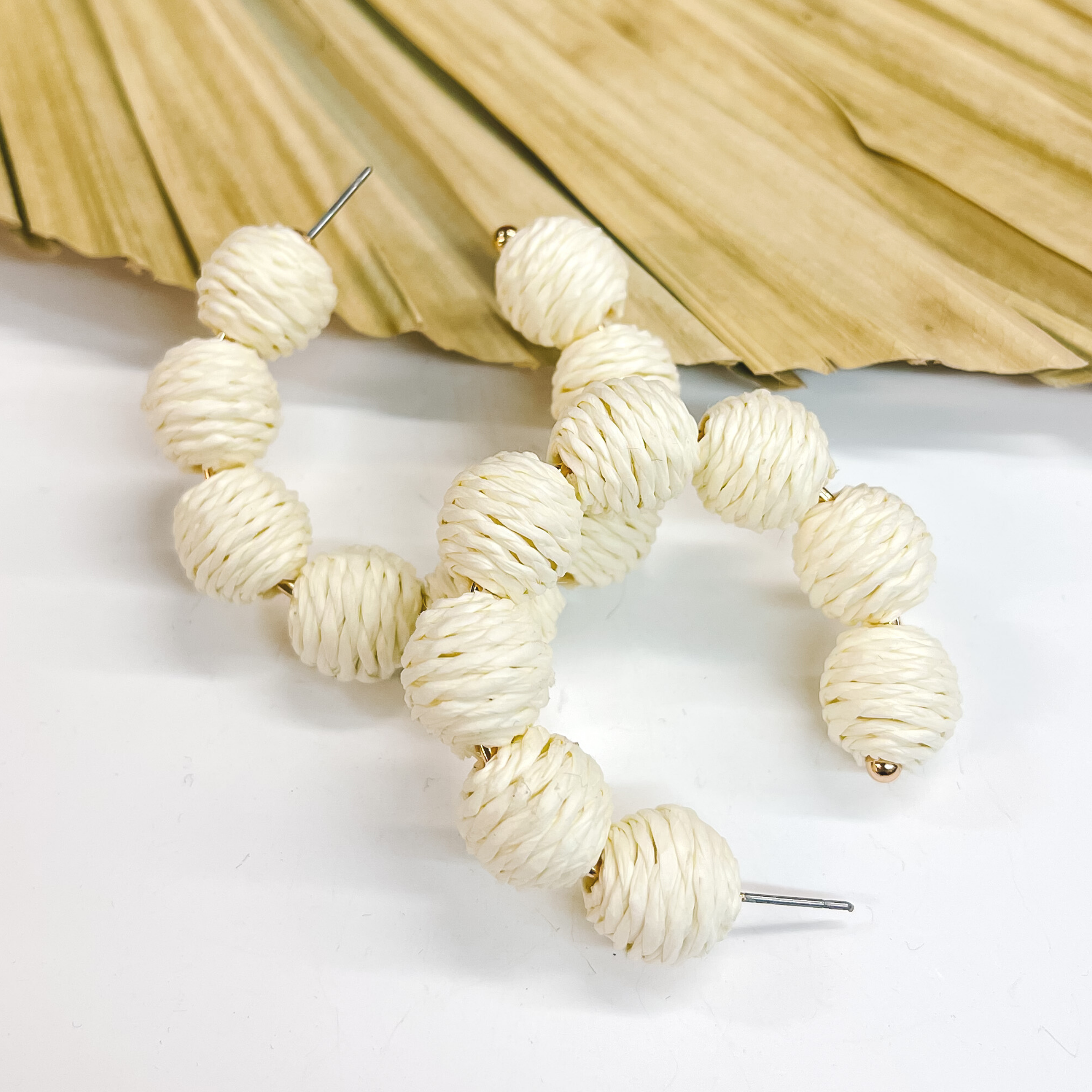 Hoop earrings with raffia balls around. The balls are wrapped in ivory raffia. Taken on a white background and  leaned up against a dried up palm leaf.