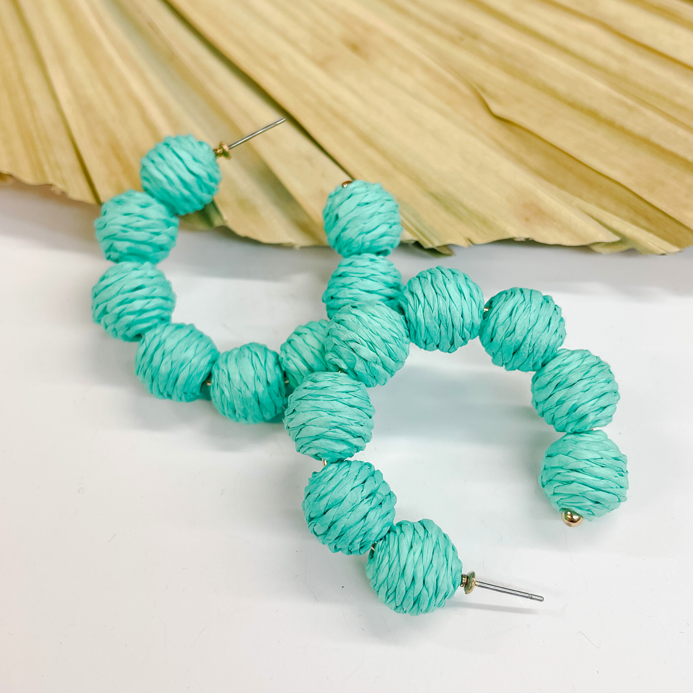 Hoop earrings with raffia balls around. The balls are wrapped in turquoise raffia. Taken on a white background and  leaned up against a dried up palm leaf.