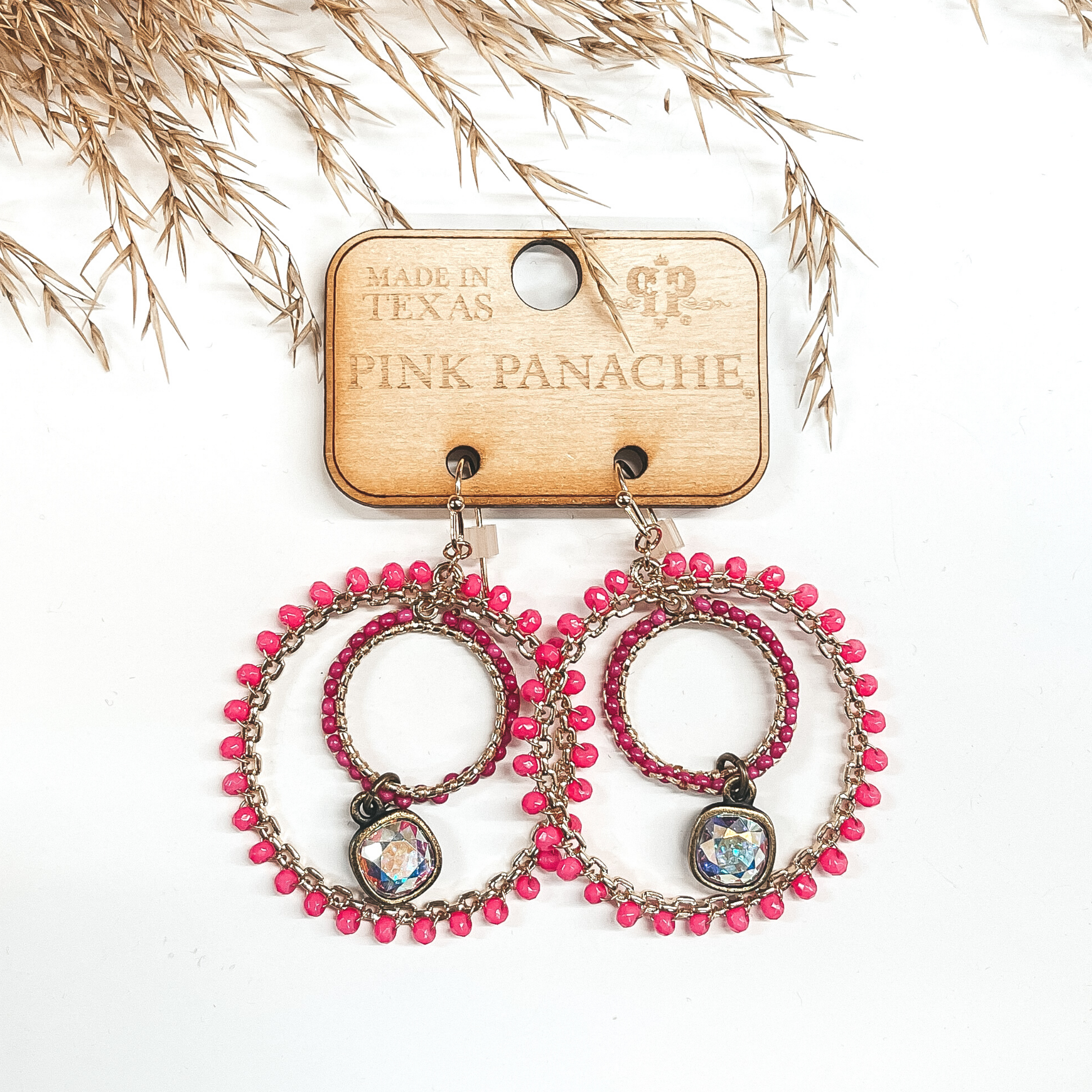 These are gold double hoop earrings with hot pink beads around on the outside hoop. The inside hoop has  smaller fuchsia beads with a hanging ab crystal in the center. Taken on a white background with a brown plant in the back as decor.