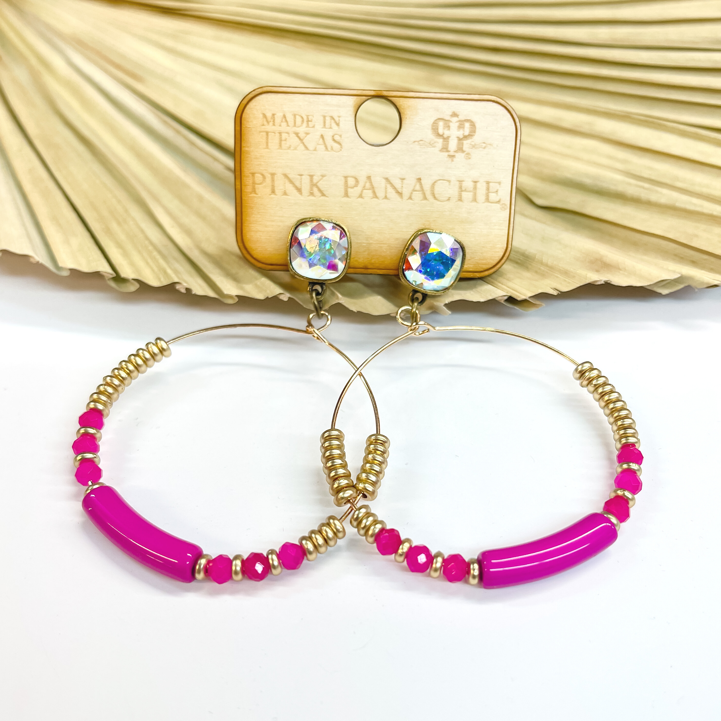 These are ab cushion cut studded earrings with a  gold circle drop. The hoop has a long fuchsia bead in  the center with matte gold beads and three fuchsia crystal beads on each side. They are taken on a white  background and leaned up against a dried up palm  leaf.
