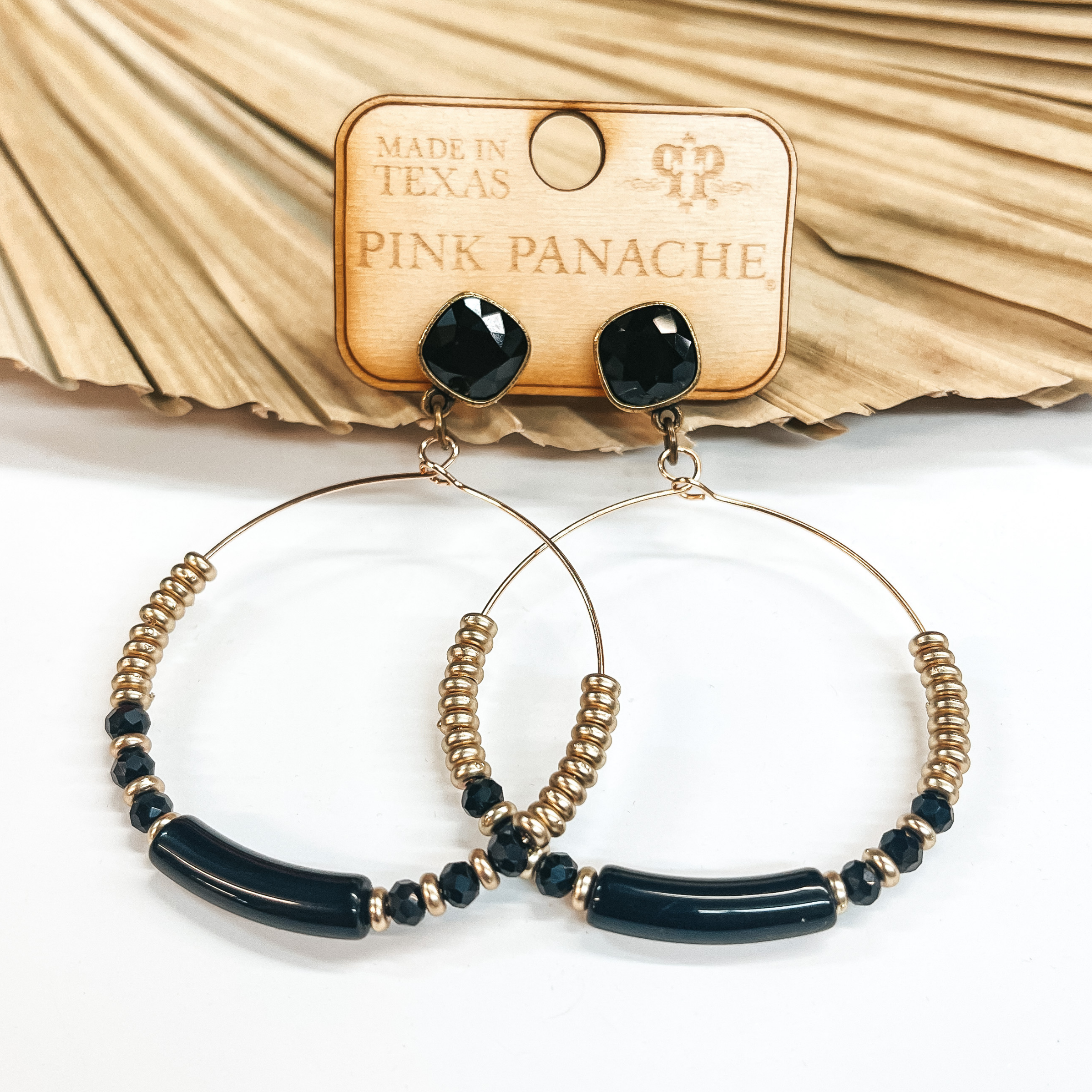 These are black cushion cut studded earrings with a  gold circle drop. The hoop has a long black bead in  the center with matte gold beads and three black crystal beads on each side. They are taken on a white  background and leaned up against a dried up palm  leaf.