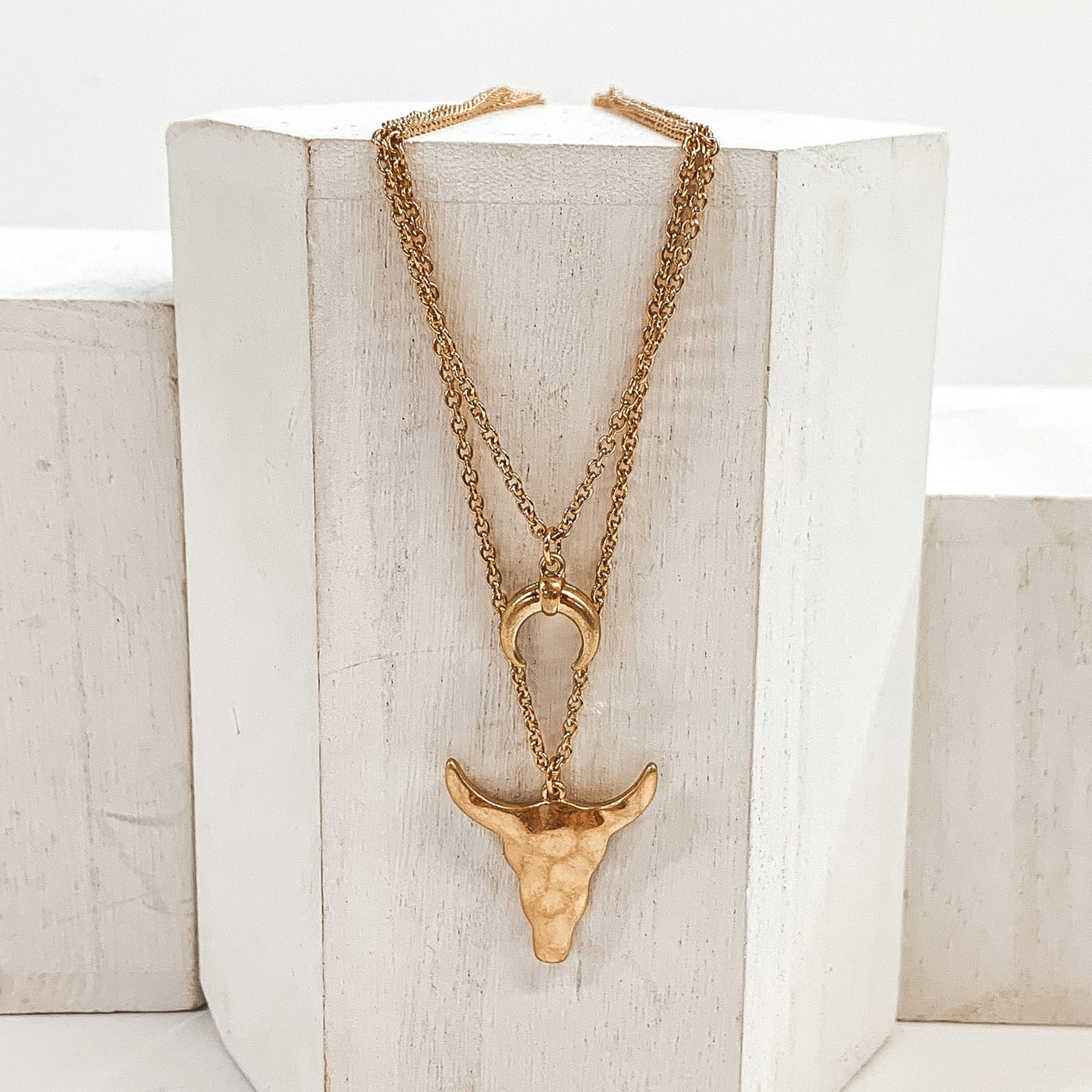 Gold double layered chained necklace with a bull skull pendant on the longer strand and horn pendant. This necklace is pictured laying on a white block on a white background.
