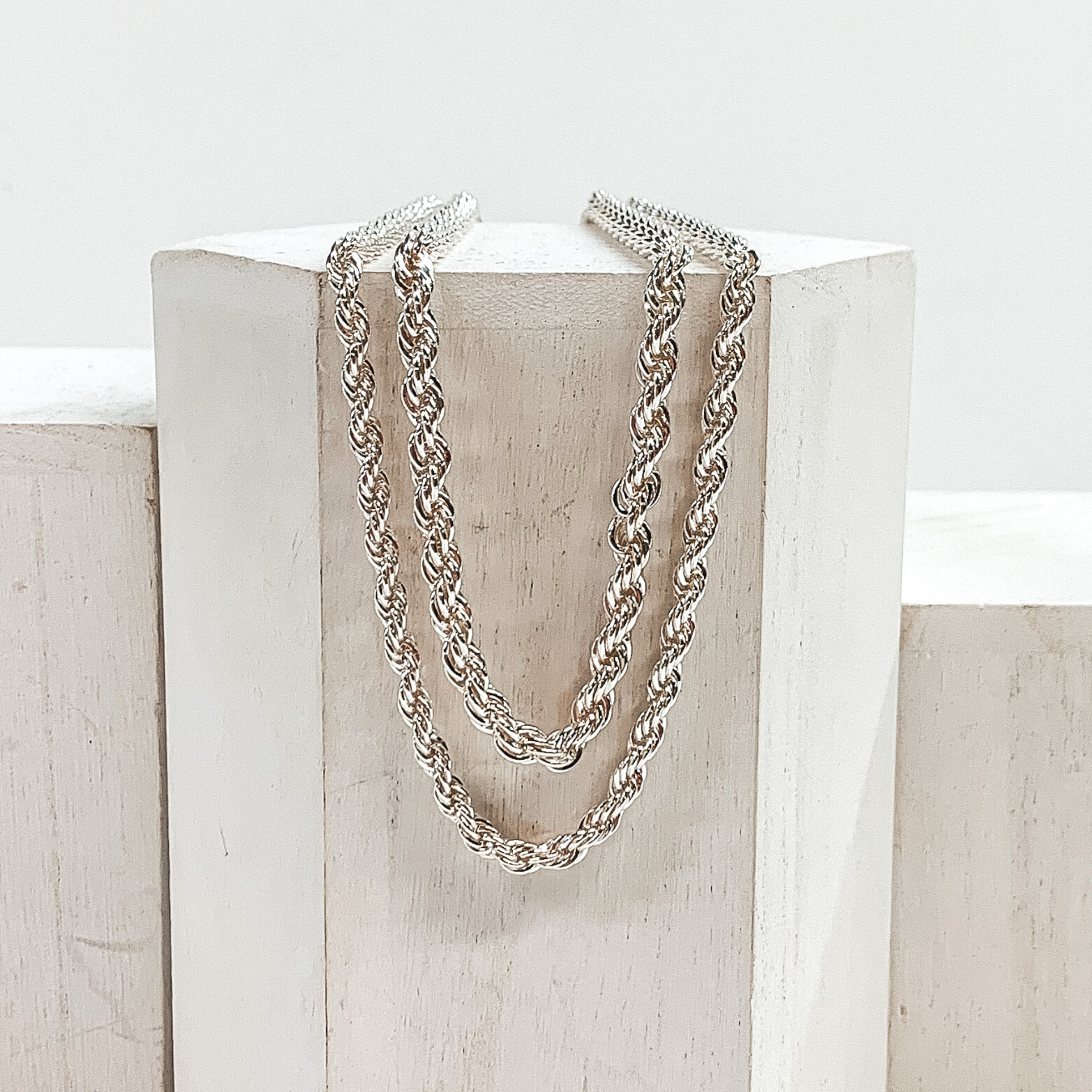 Two, silver, rope chain necklaces. This set includes a shorter and a longer necklace. This necklace is pictured laying on a white block on a white background.