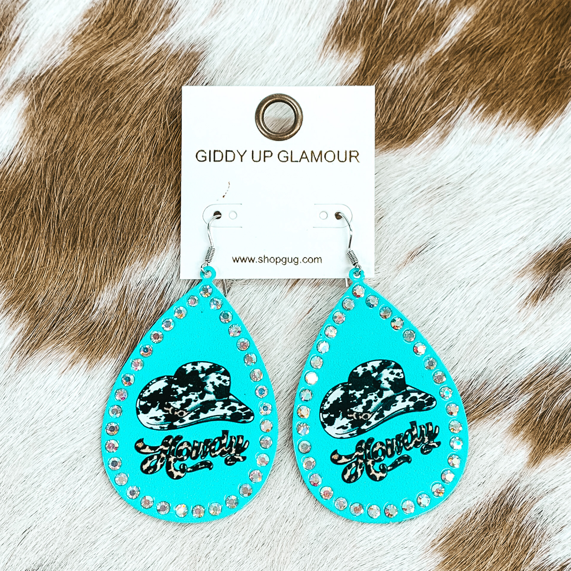 Light blue teardrop earrings with an AB crystal outline. In the center of the earrings, there is a black and white cow print cowboy hat with a leopard print band. There is also the word "Howdy in cursive underneath the cowboy hat in a leopard print. These earrings are pictured on a brown and white cow print hide material.