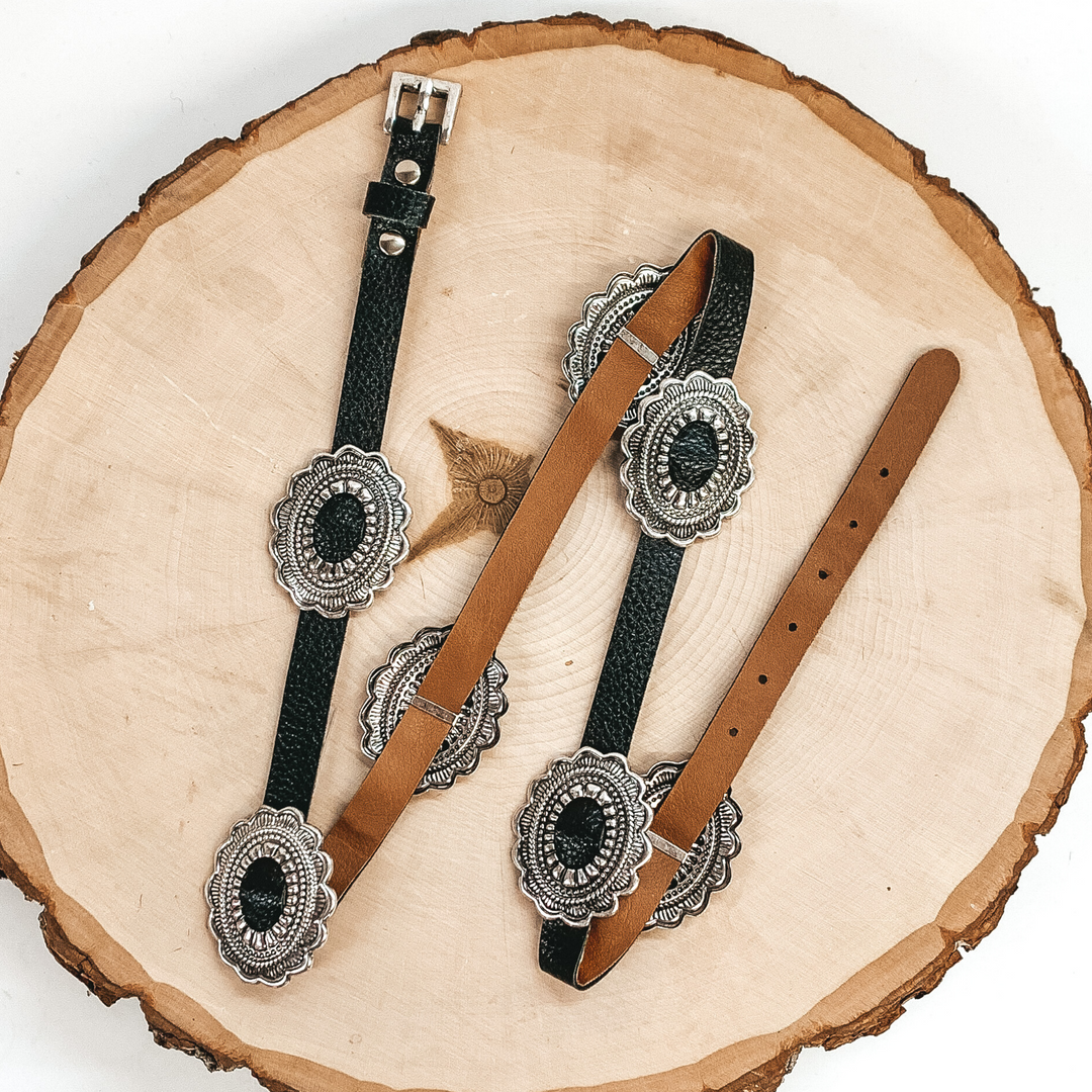 Black hat band with silver, spaced out conchos. This hat band is pictured laying on a piece of wood on a white background.