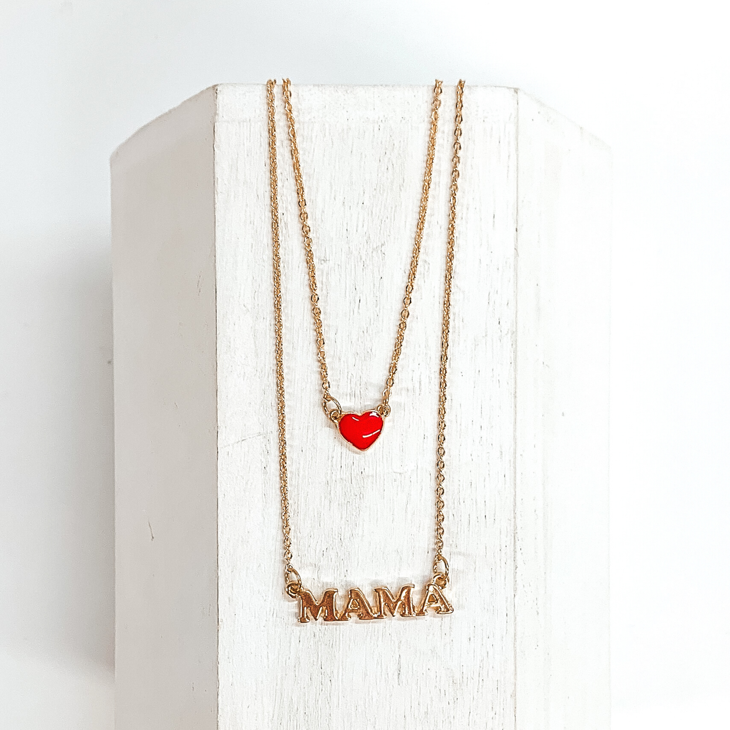 Two stranded gold chained necklace. The longer strand has a gold pendant that spells out "MAMA" and the shorter strand has a small red heart charm. This necklace is pictured laying on a white block on a white background. 