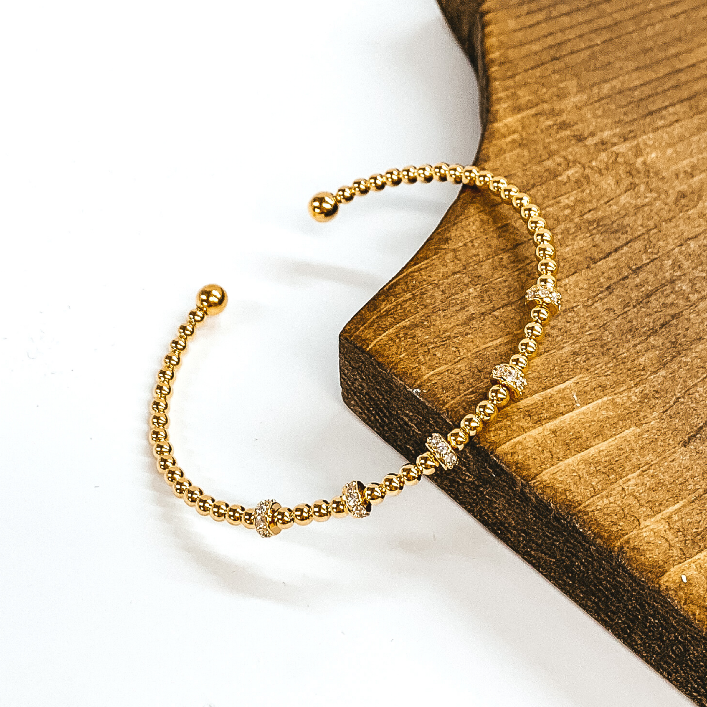 Gold beaded bangle bracelet with clear crystals spacers. This bangle is pictured leaning on a brown block on a white background.