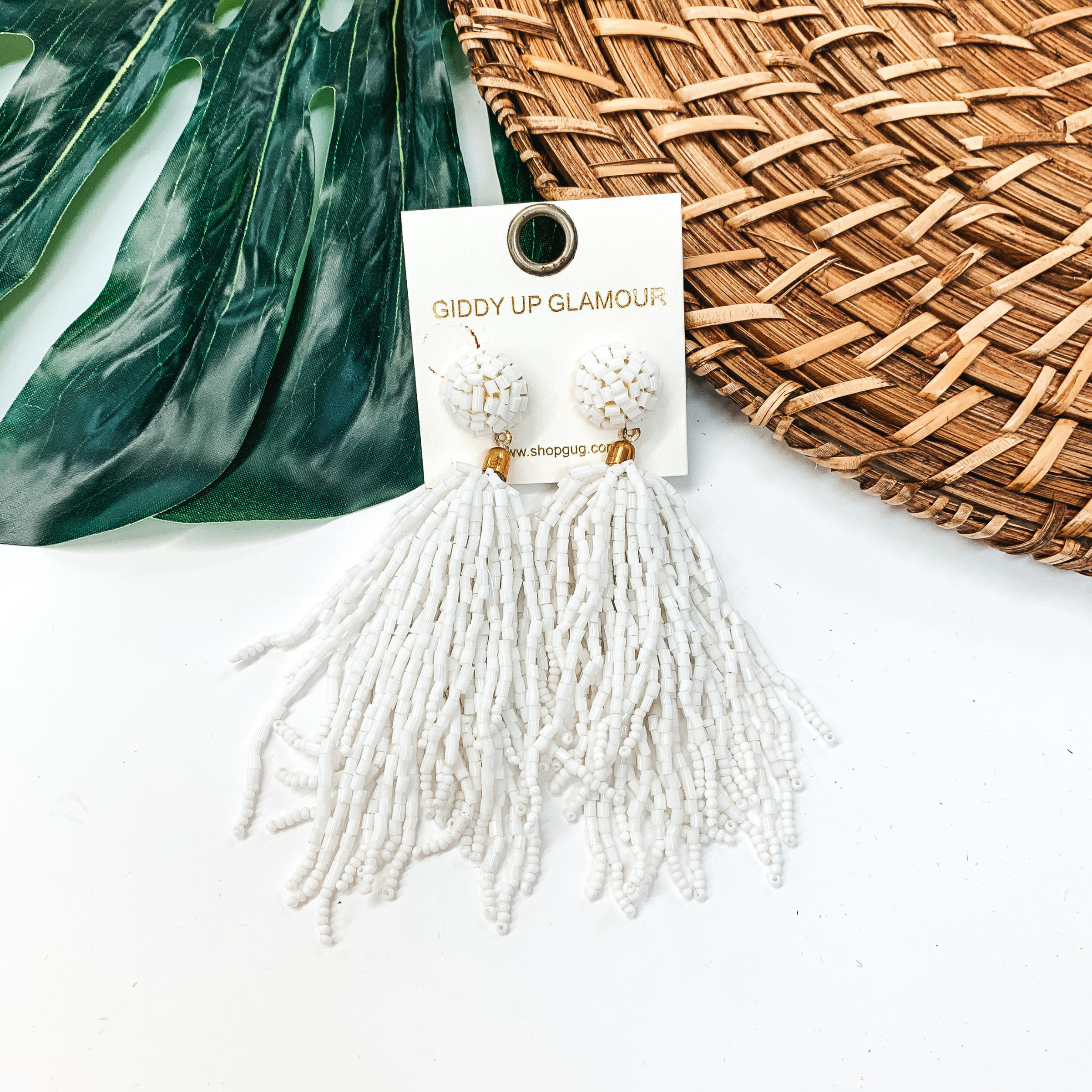 Circle, beaded post back stud earrings with a hanging beaded tassel in white. These earrings are pictured on a white background with a green leaf and tan basket weave in the background.