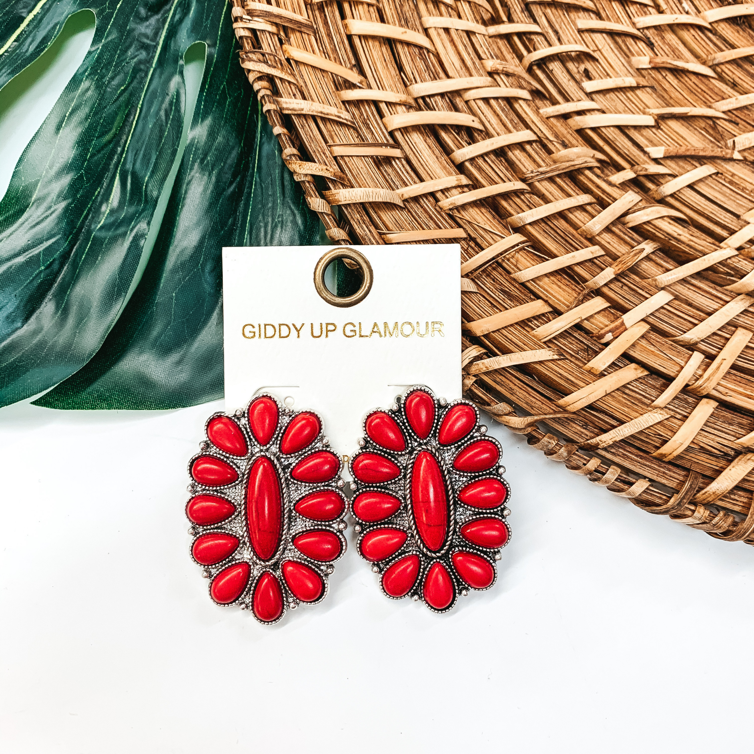 Oval cluster earrings with red stones. These earrings are pictured on a white background in front of a green leaf. 