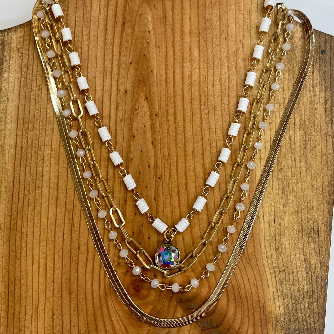 Gold four strand necklace with an AB cushion cut crystal pendant. The outer strand is a snake chain followed by a gold chain with ivory beads all around. The third strand is a matte gold link chain. The  smaller strand is a gold chain with white disks  all around and an AB cushion cut crystal pendant.  Taken on a brown necklace block and white background.