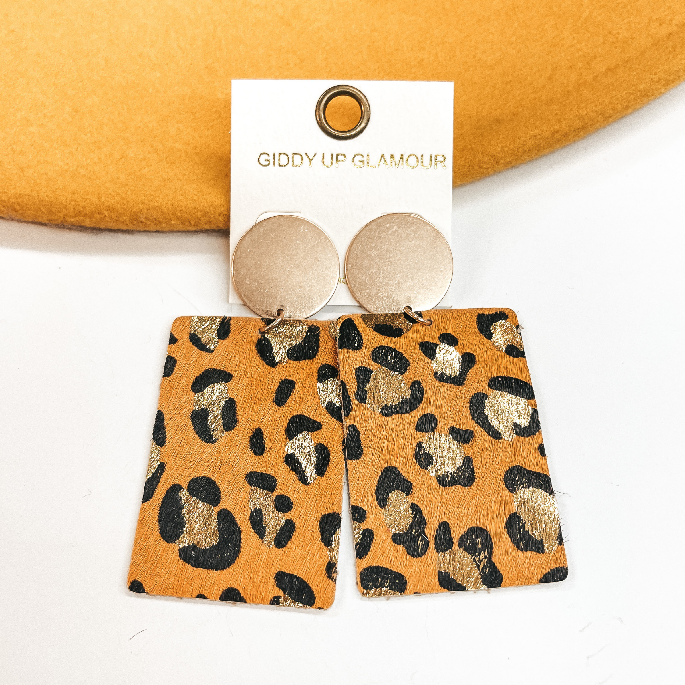 Gold circle stud earrings with black and gold leopard print on yellow leather hide.  Taken on a white background and  leaned up against a yellow hat.