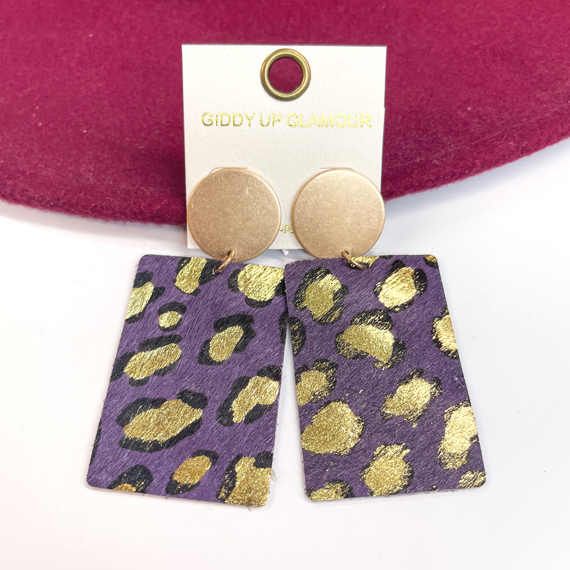 Gold circle stud earrings with black and gold leopard print on dark purple leather hide.  Taken on a white background and  leaned up against a burgundy hat.