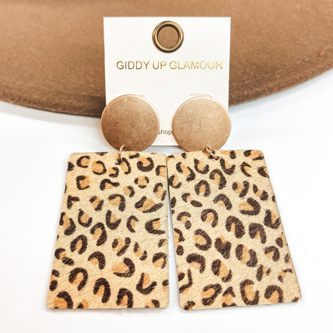 Gold circle stud earrings with dark and light brown leopard print on beige leather hide.  Taken on a white background and  leaned up against a light brown hat.