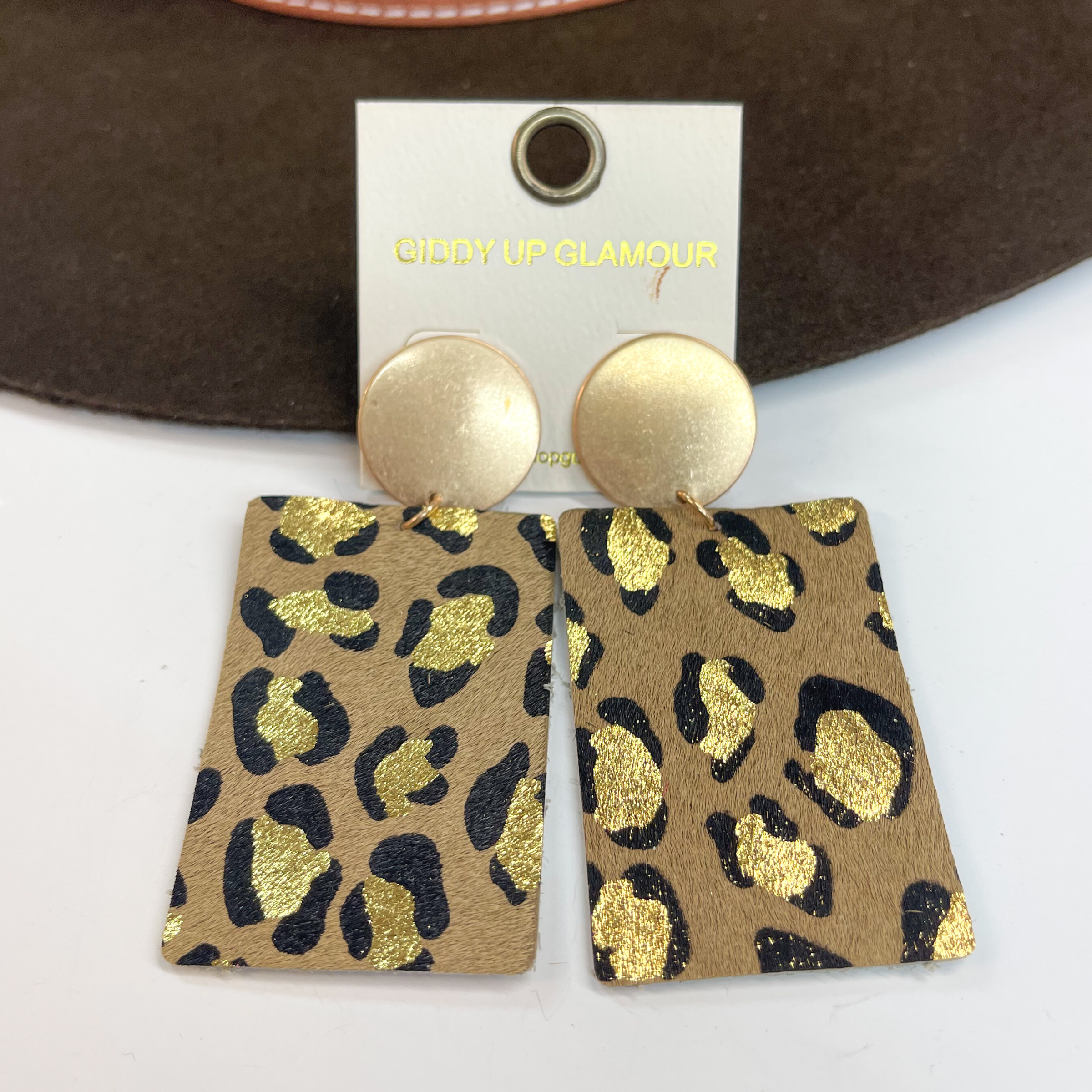 Gold circle stud earrings with black and gold  leopard print on dark brown leather hide.  Taken on a white background and  leaned up against a dark brown hat.