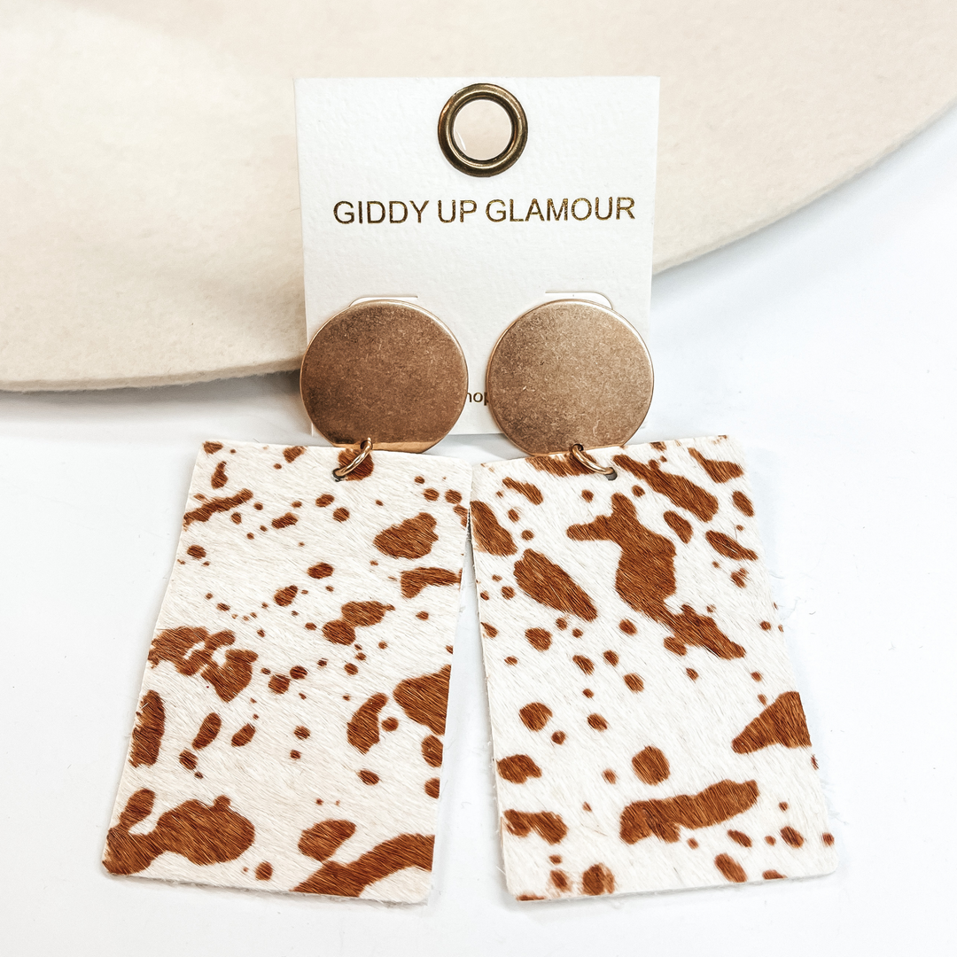 Gold circle stud earrings with brown cow  print on white color leather hide.  Taken on a white background and  leaned up against an ivory hat.