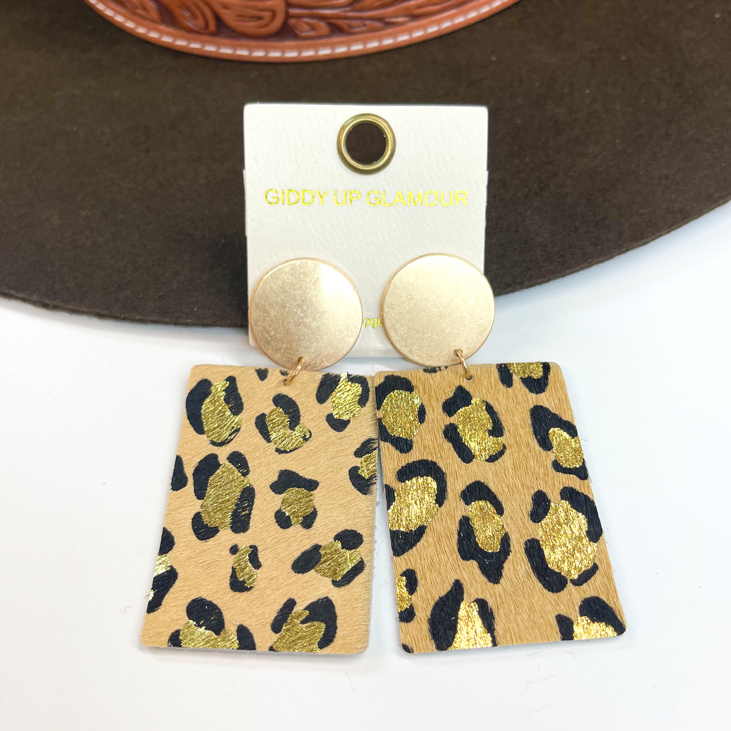 Gold circle stud earrings with black and gold  leopard print on beige color leather hide.  Taken on a white background and  leaned up against a dark brown hat.