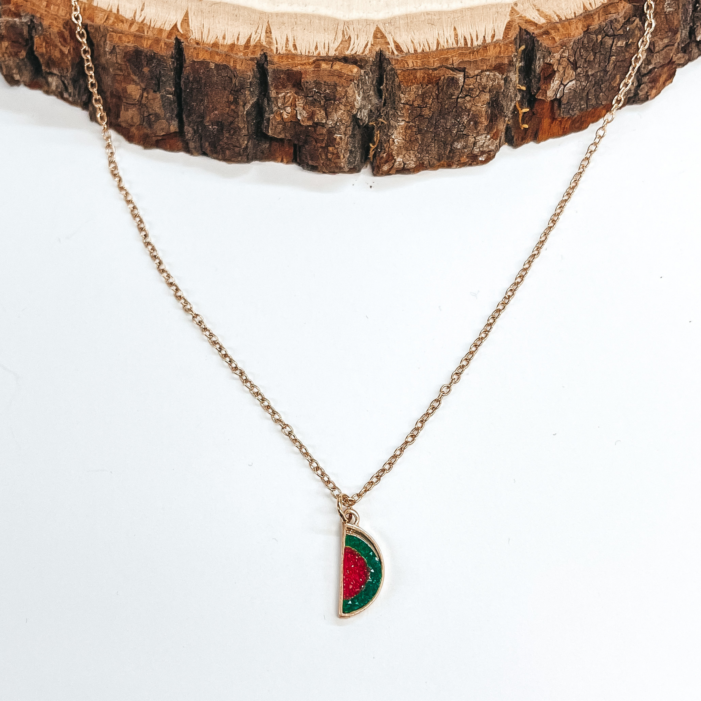 Gold necklace with a watermelon druzy pendant in  green and red. Taken on a white background and a  piece of wood. 