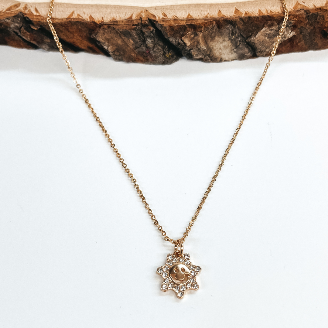 Gold necklace with sun shaped pendant. The sun shaped pendant has clear crystals all around and a happy  face in the center. Taken on a white background and a piece of wood.