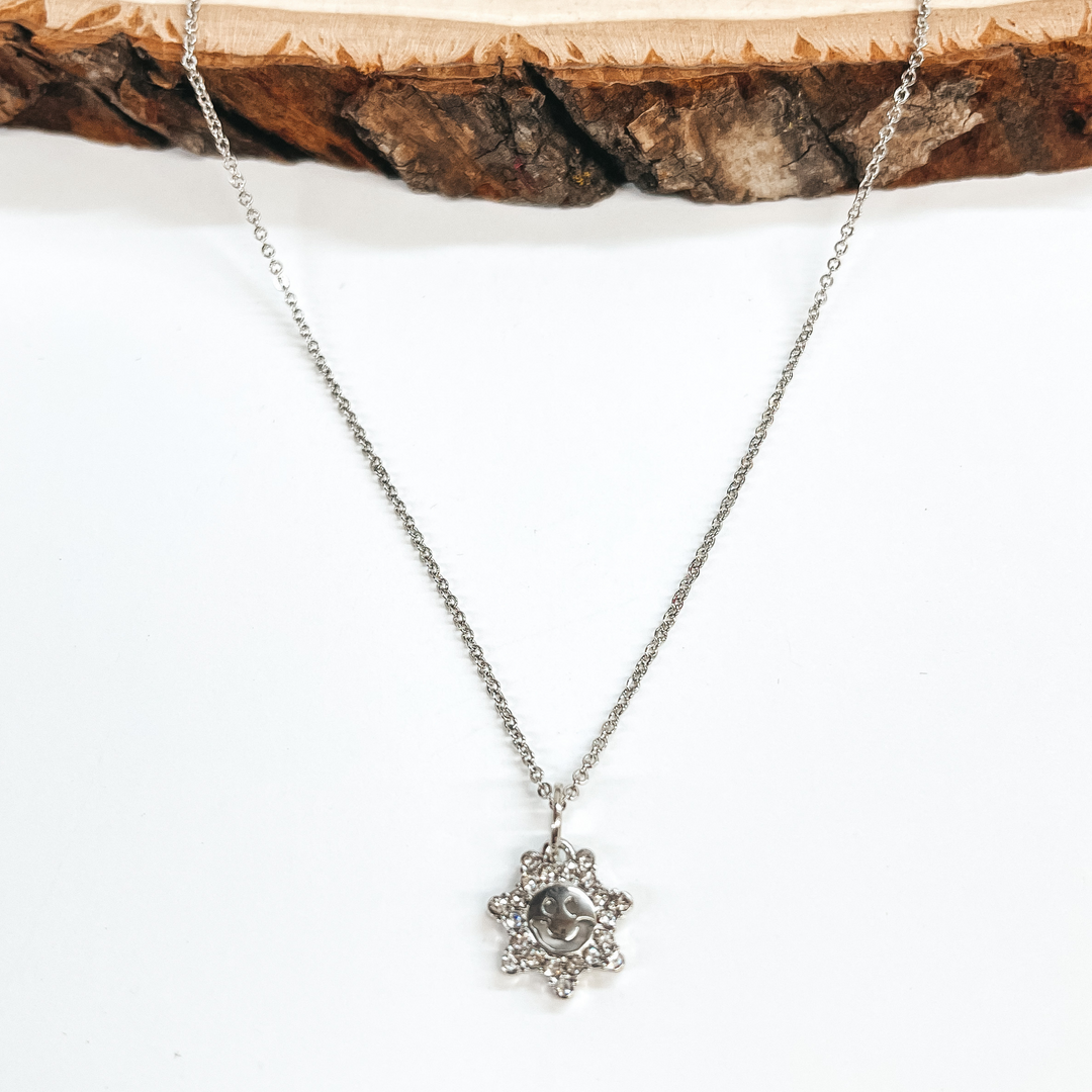 Silver necklace with sun shaped pendant. The sun  shaped pendant has clear crystals all around and a happy  face in the center. Taken on a white background and a piece of wood.
