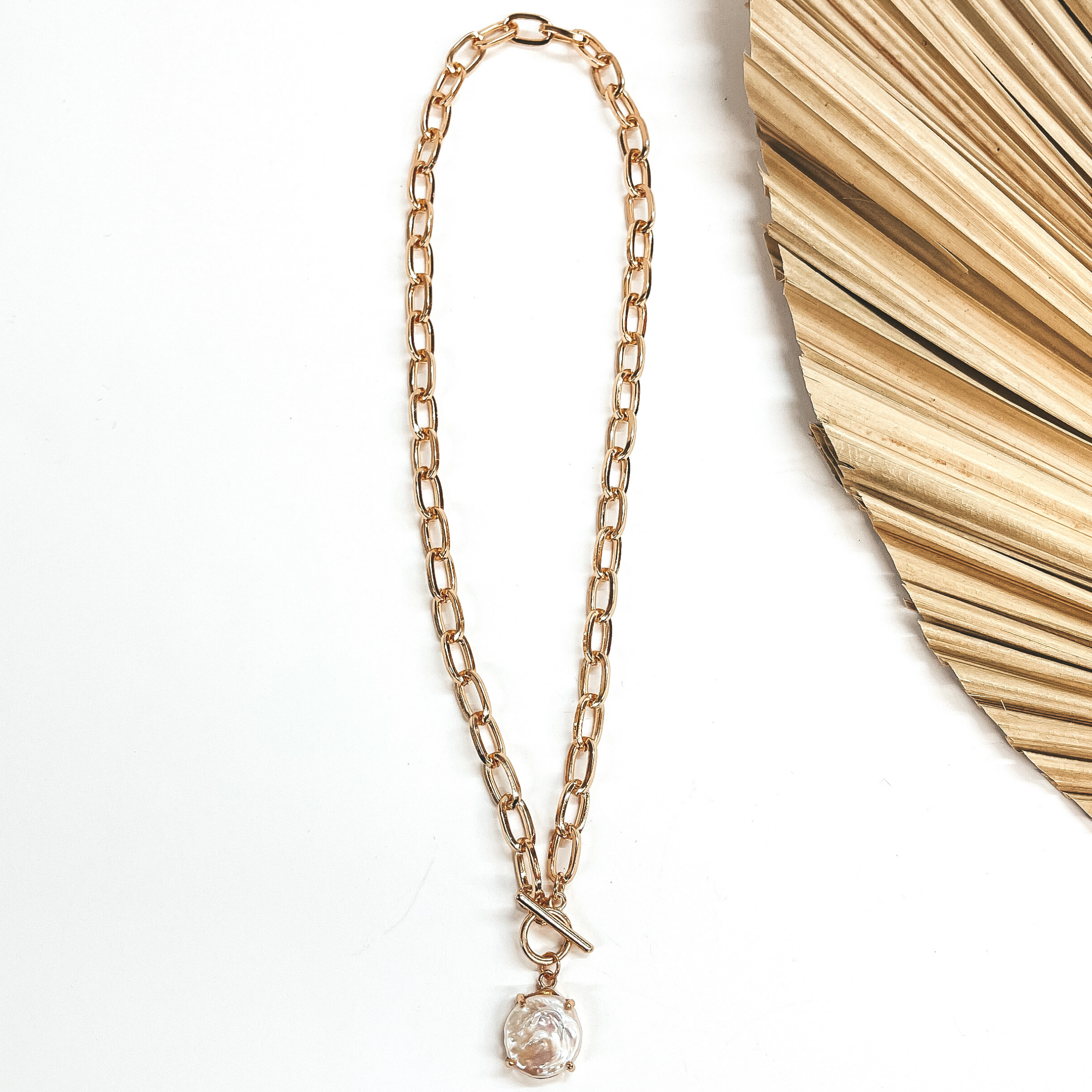 Gold thick chain with a toggle clasp and  freshwater pearl pendant. Taken on a white background and a dried up palm leaf in the side as decor.