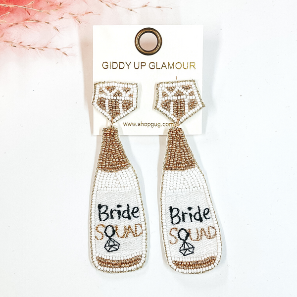 Post back earrings with a bottle drop in white and gold beads. The stud part has a diamond shape in beads, the bottle has the saying 'Bride Squad' with a diamond ring replacing the Q. Taken on a white  background with a pink plant in the back as decor.