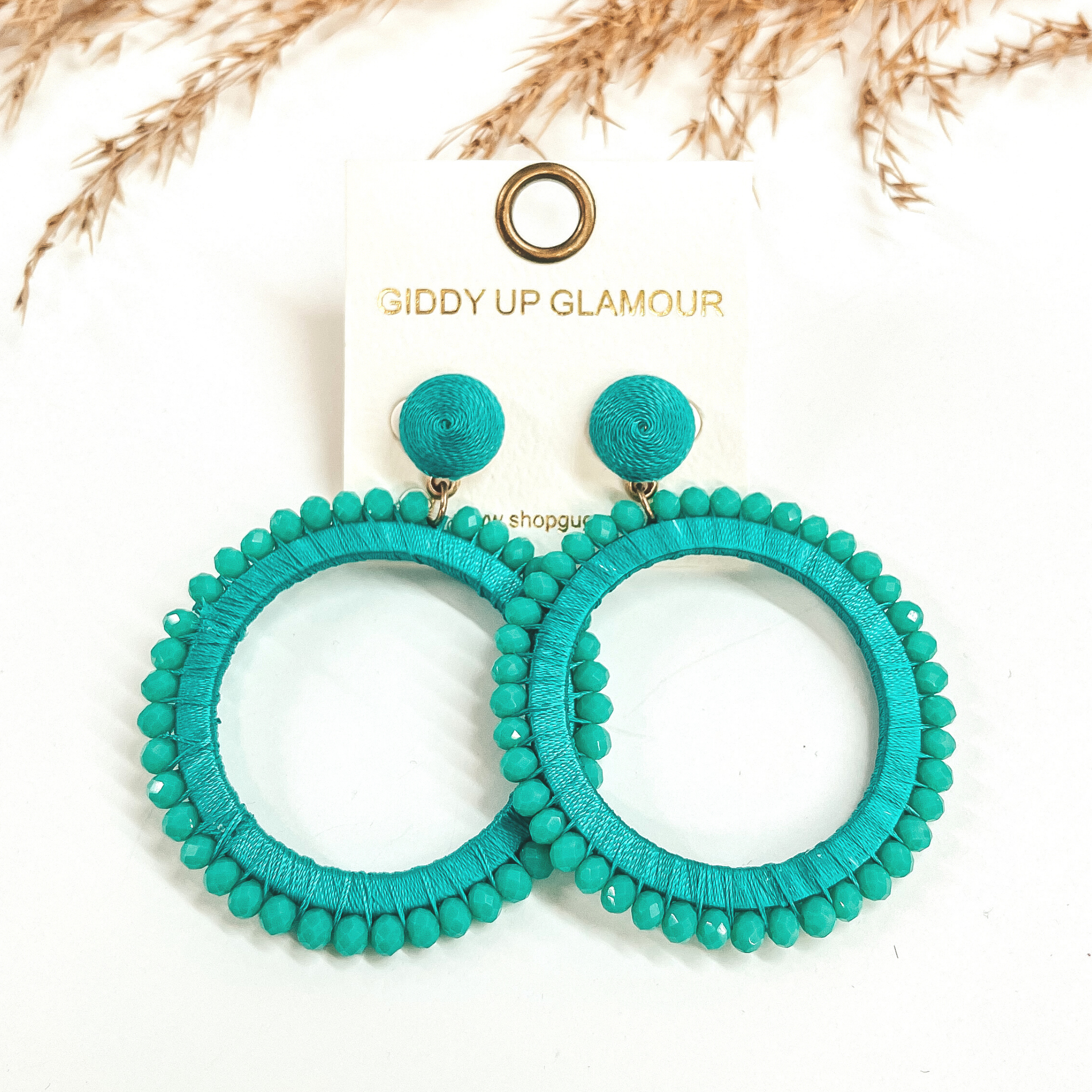 Turquoise post back circle drop earrings. Turquoise colored beads all around the circle and the circle is wrapped with turquoise colored thread.  Taken on a white background with a brown plant in the back as decor.