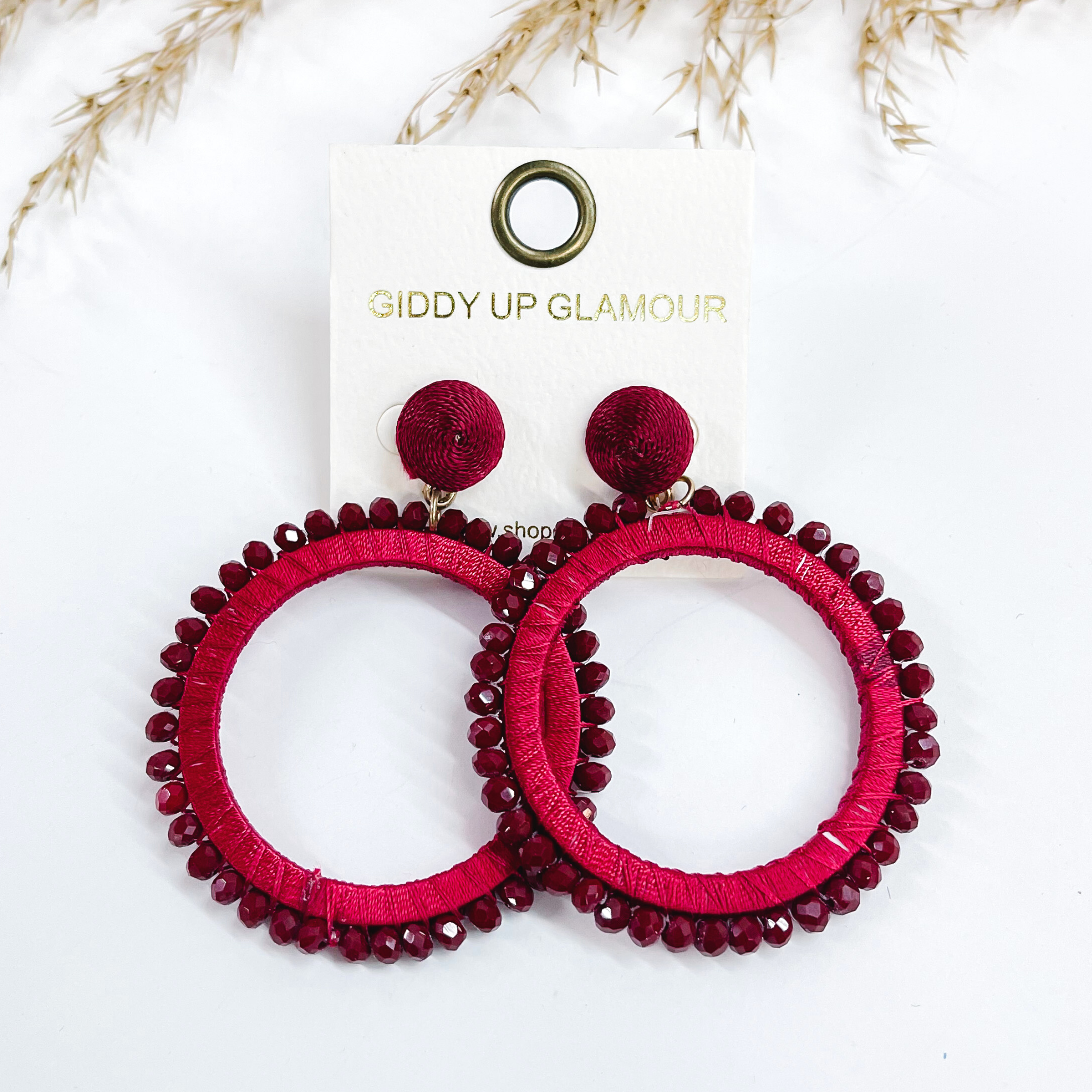 Burgundy post back circle drop earrings. Burgundy colored beads all around the circle and the circle is wrapped with burgundy colored thread.  Taken on a white background with a brown plant in the back as decor.