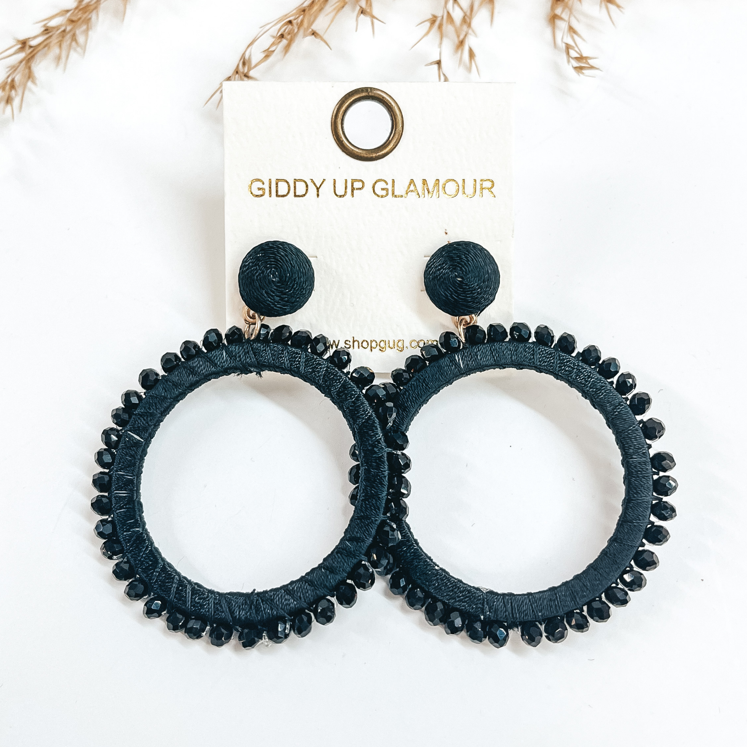 Black post back circle drop earrings. Black colored beads all around the circle and the circle is wrapped with black colored thread.  Taken on a white background with a brown plant in the back as decor.