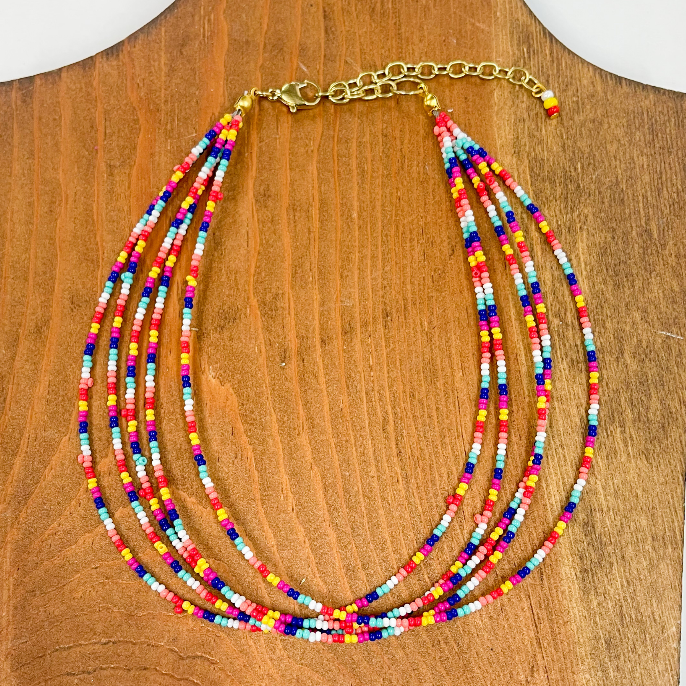 Multilayered seedbeaded choker in multicolor.  The multicolored beads come in white, coral,  orange, red, yellow, dark blue, turquoise, and hot  pink. The adjustable  chain has three beads in colors, white, yellow, and  red. These earrings are taken on a brown block.