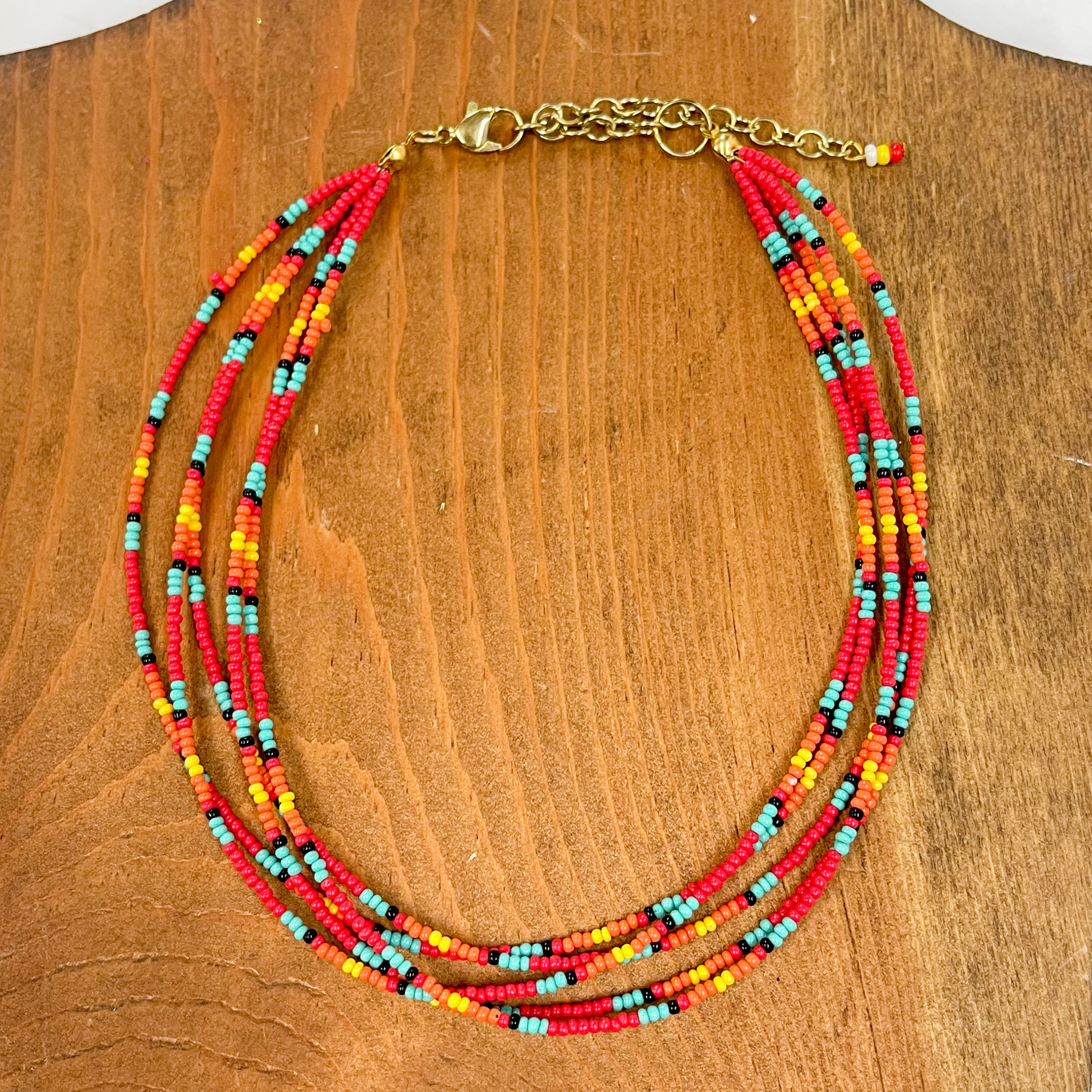 Multilayered seedbeaded choker in red multicolor.  Majority of the beads are red, other colors include; turquoise, black, orange, and yellow. The adjustable  chain has three beads in colors, white, yellow, and  red. These earrings are taken on a brown block.