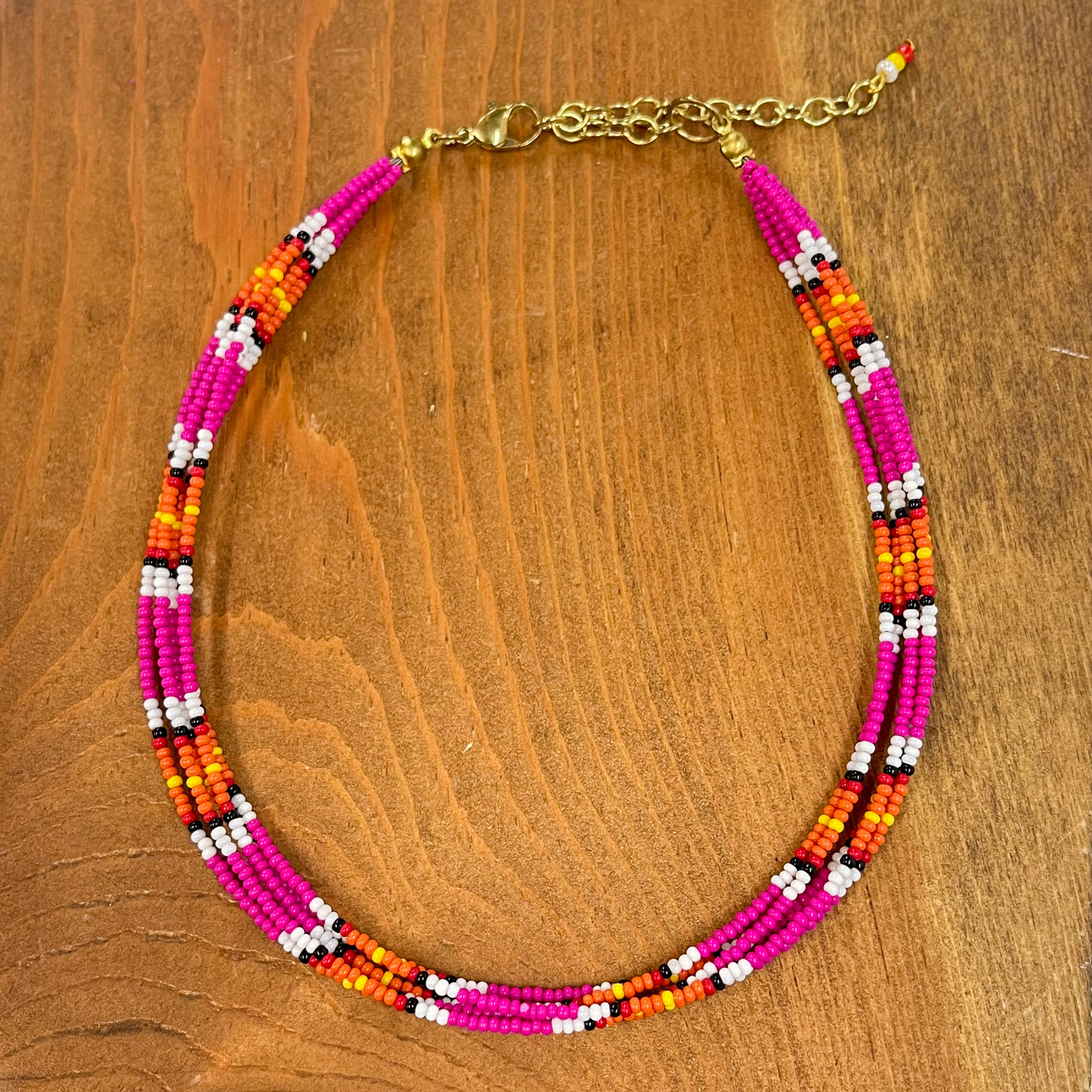Multilayered seedbeaded choker in pink multicolor.  Majority of the beads are pink, other colors include; white, black, orange, red, and yellow. The adjustable  chain has three beads in colors, white, yellow, and  red. These earrings are taken on a brown block.