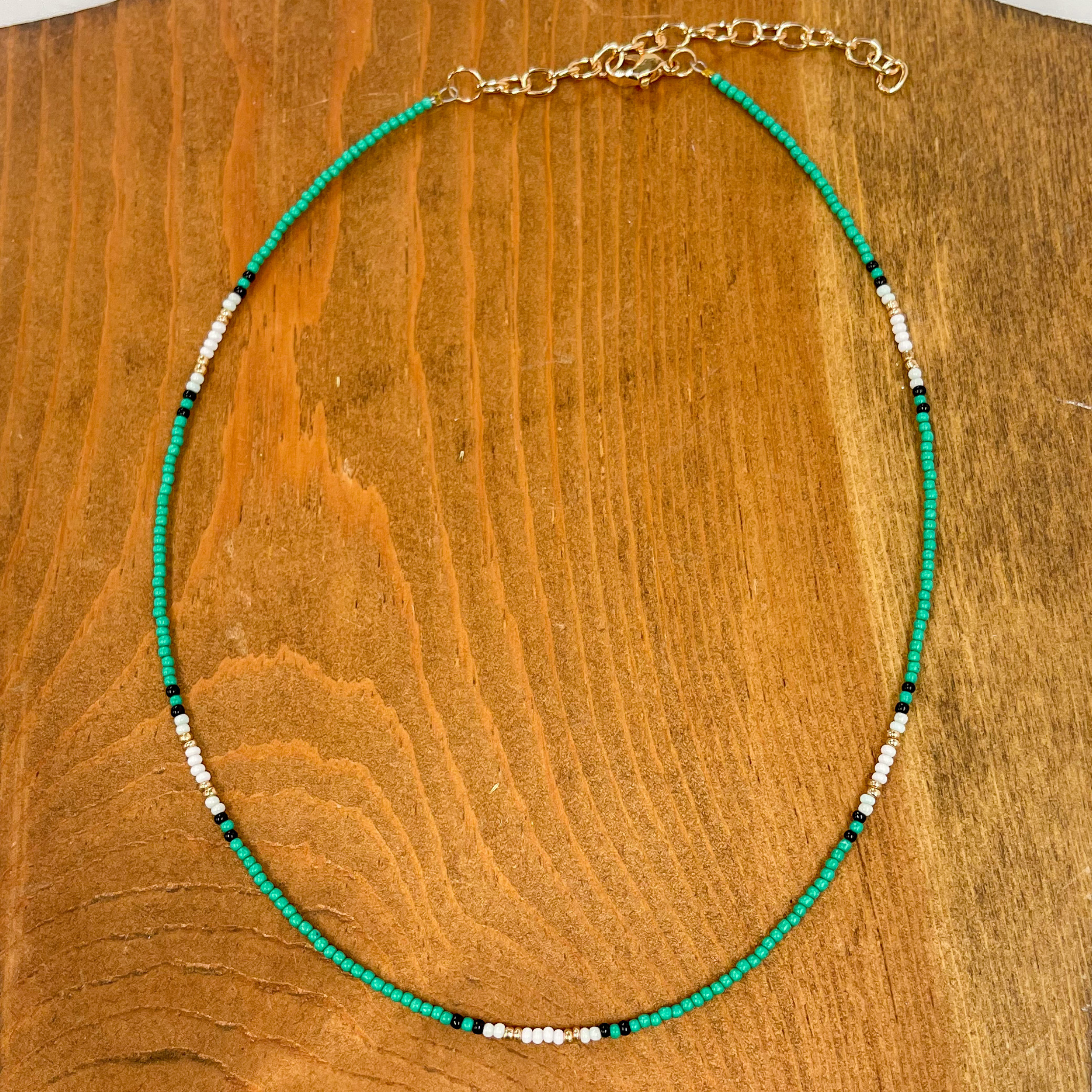 Seed beaded necklace with a gold adjustable. The  majority of the beads are teal with a few black, white,  and gold beads. This necklace is taken on a brown  block.