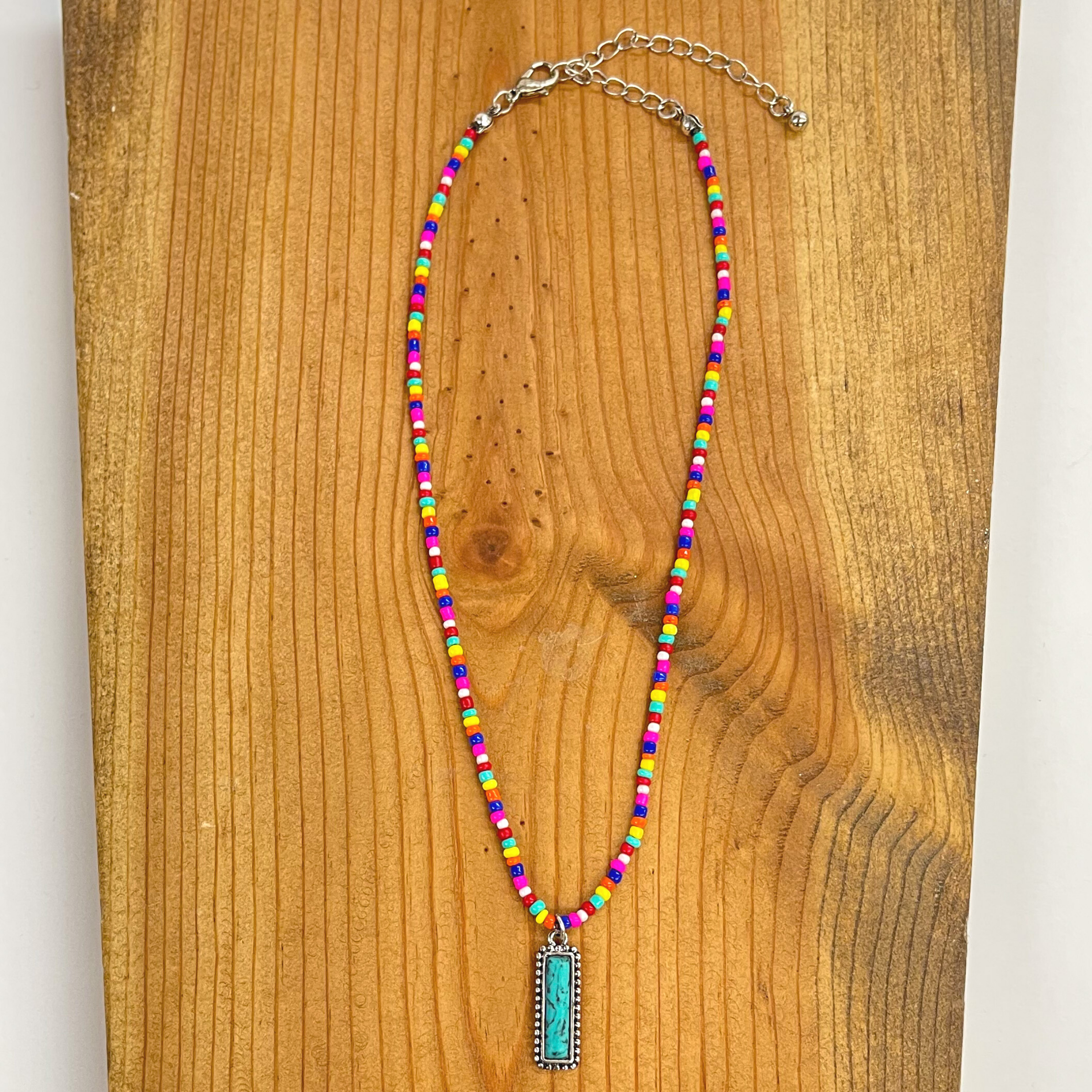 Multicolored seed beaded necklace with a silver  adjustable and pendant. The beads  are turquoise, hot pink, dark blue, red, orange,  yellow, and white. There is a turquoise stone bar  pendant in the middle in a silver setting. This necklace is taken on a brown  block.