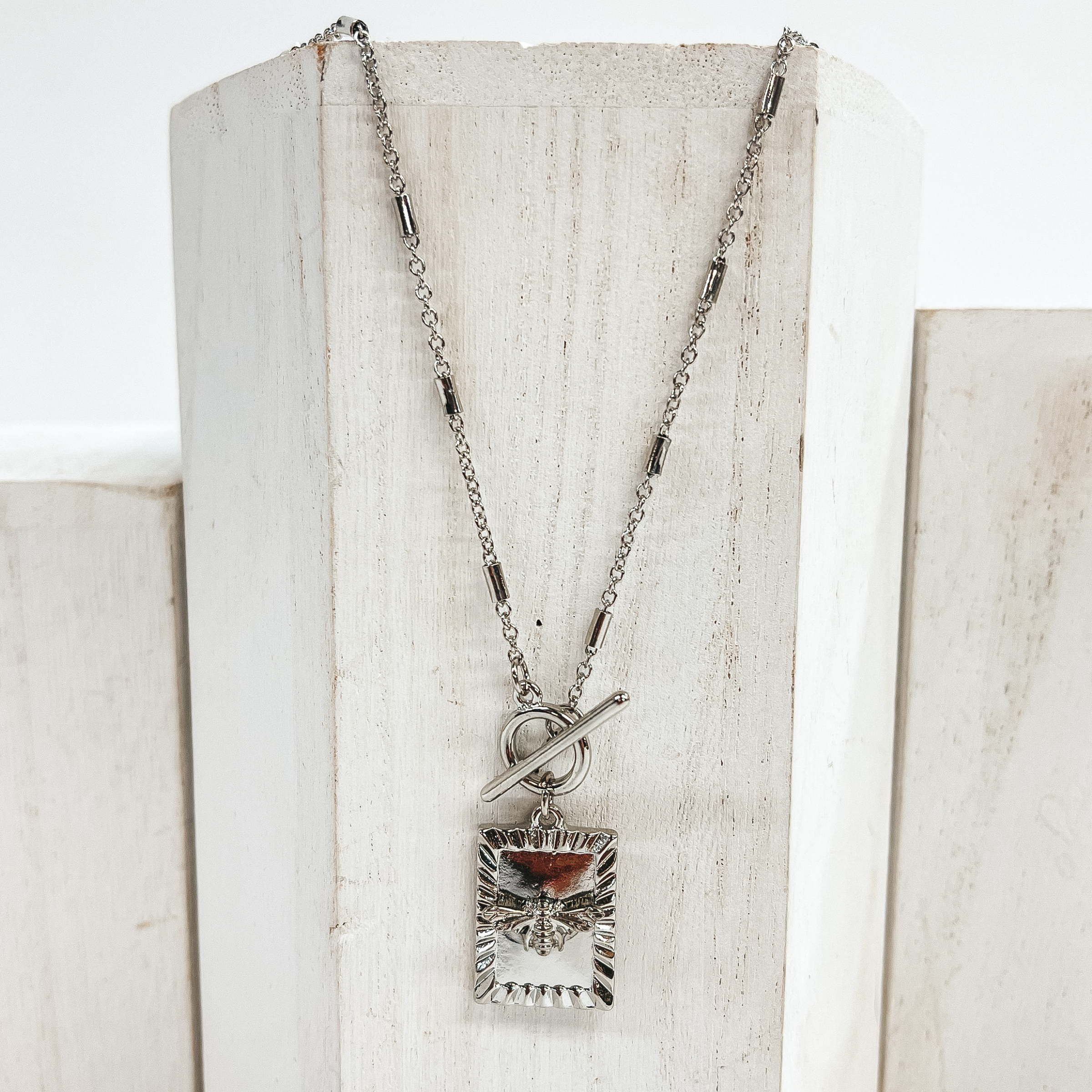 This is a silver necklace with silver spacers along  the chain. It has a front toggle clasp and a  square pendant with a bumble  bee charm. This necklace is pictured laying on a  white block and on a white background