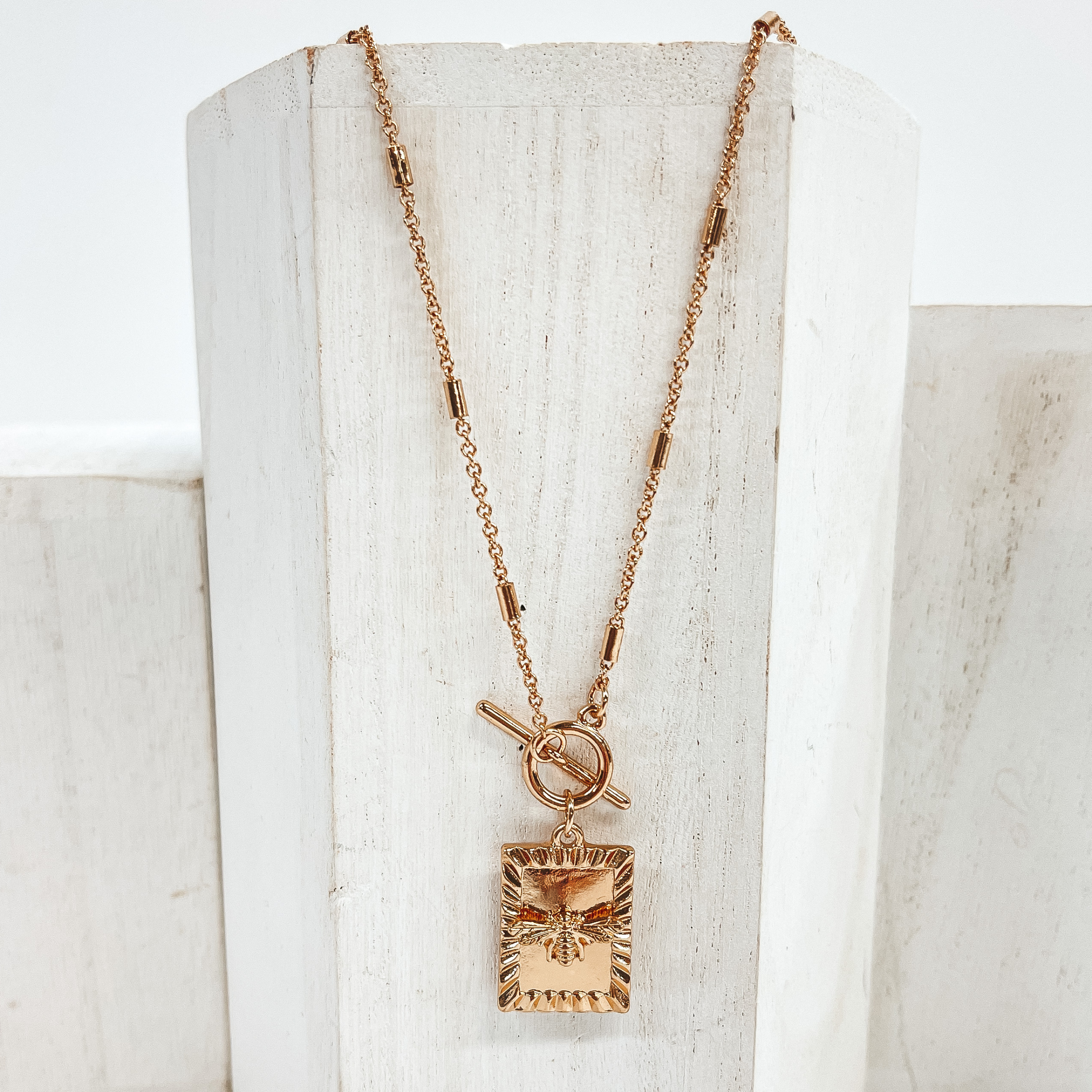 This is a gold necklace with gold spacers along  the chain. It has a front toggle clasp and a  square pendant with a bumble  bee charm. This necklace is pictured laying on a  white block and on a white background