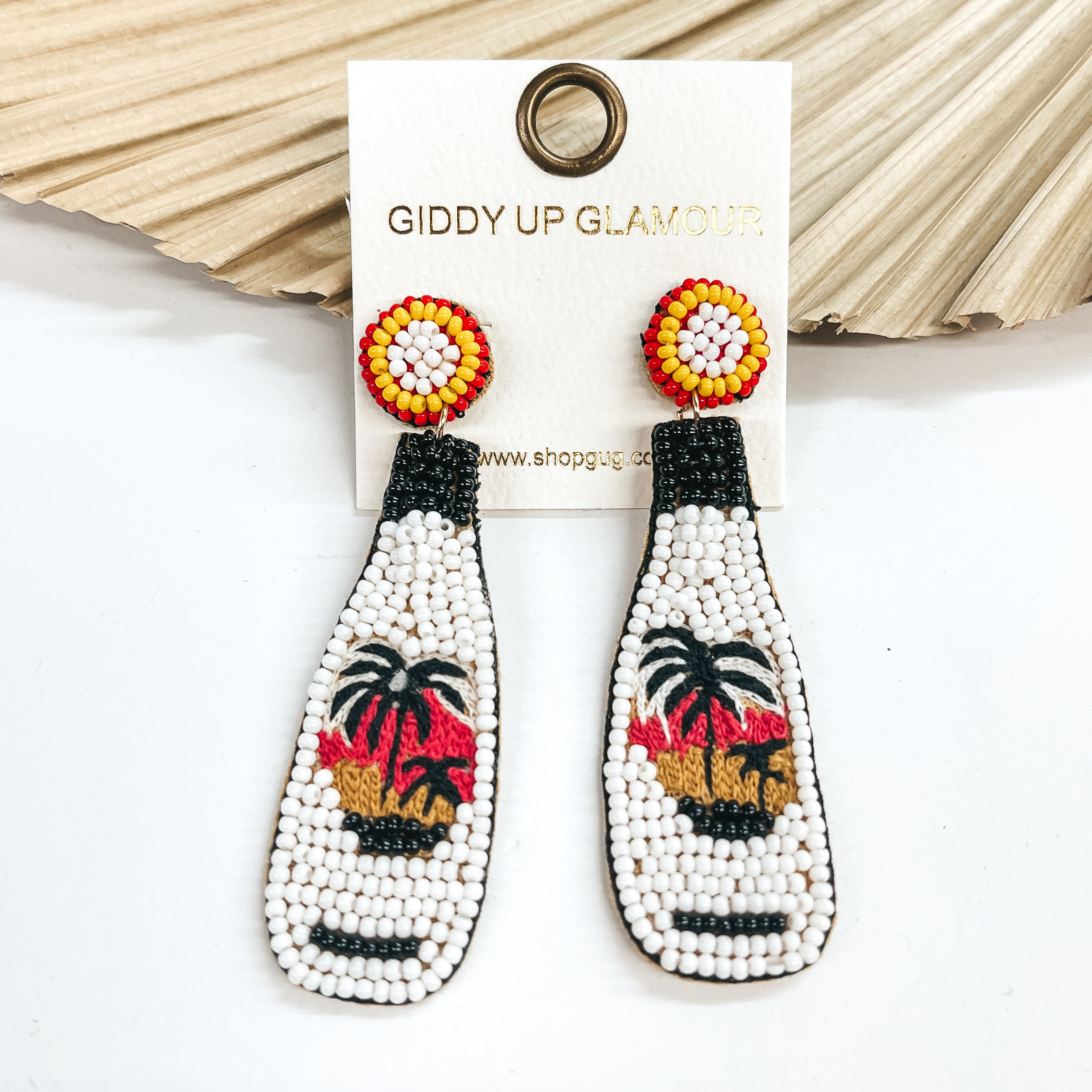 Post back earrings with a bottle drop with white, black, yellow, and red beads. In the middle there are  embroidered palm trees in a sunset. Taken on a white background and leaned up against a dried palm leaf.