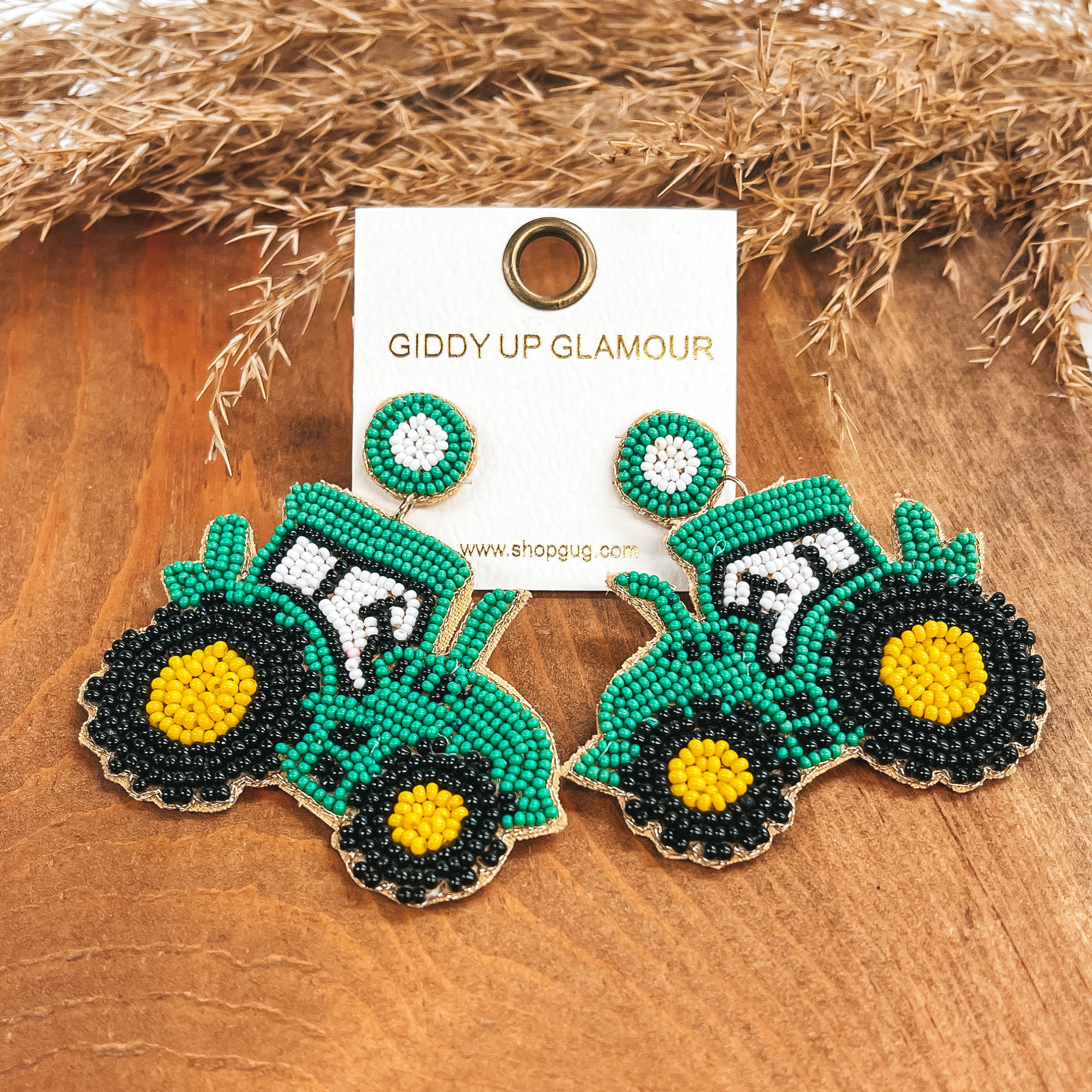 Post back earrings with a green tractor drop with green, yellow, black, and white beads. Taken on a  brown block with a brown plant in the back as decor.