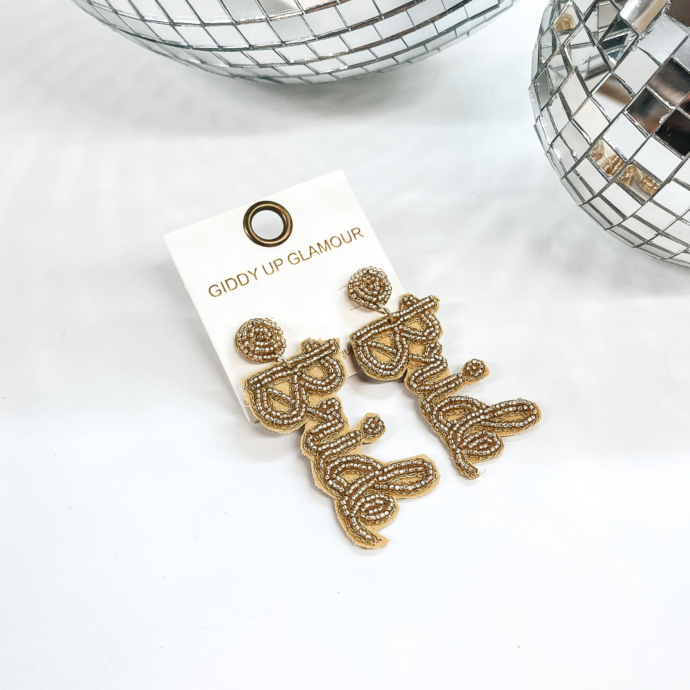 Gold beaded post back earrings with the word 'Bride' in Barbie font. Takn on a white background with two  disco balls in the back as decor.