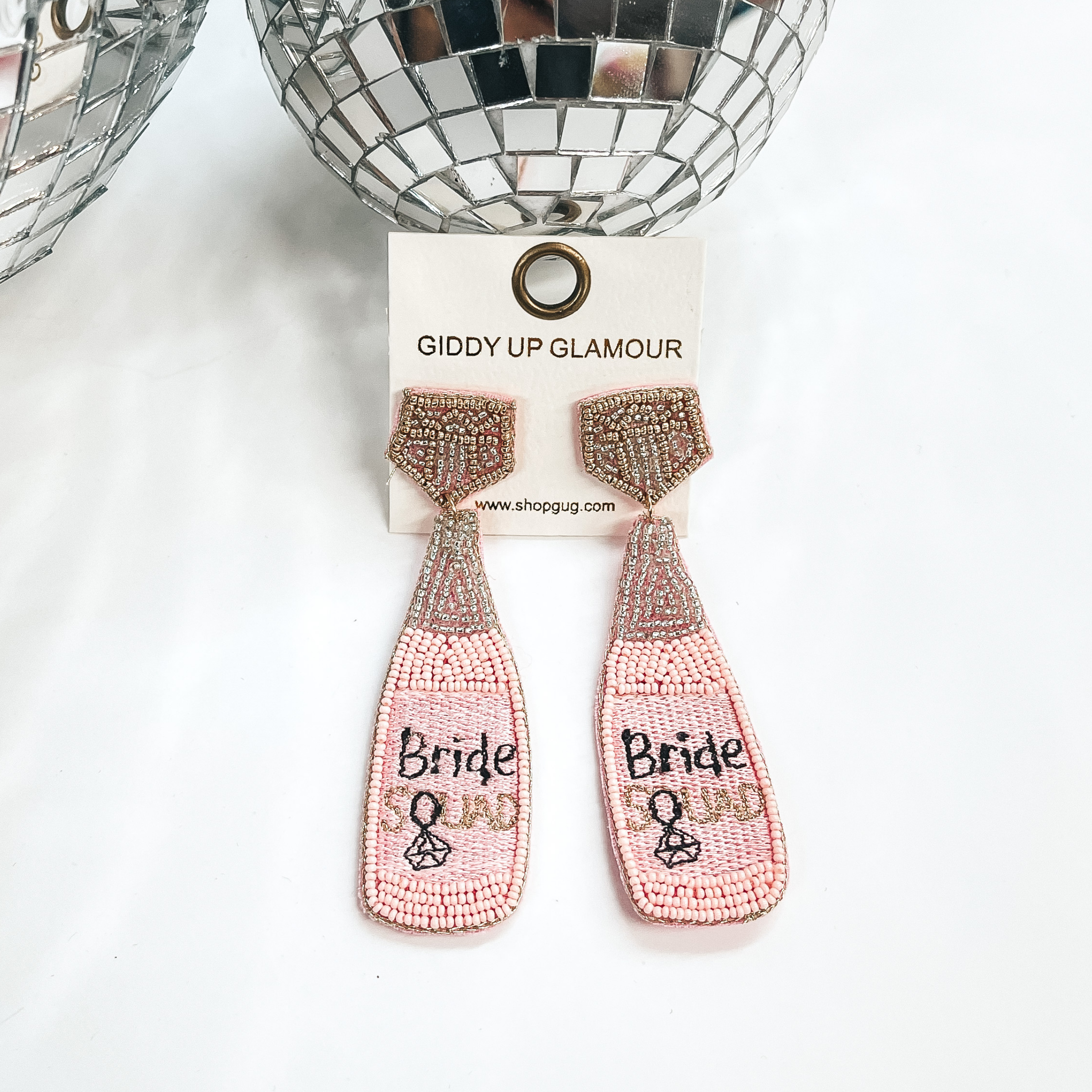 Post back earrings with a bottle drop in light pink,  gold, and silver beads. The stud part has a  diamond shape in beads, the bottle has the saying  'Bride Squad' with a diamond ring replacing the Q.  Taken on a white background with two disco balls in  the back as decor.