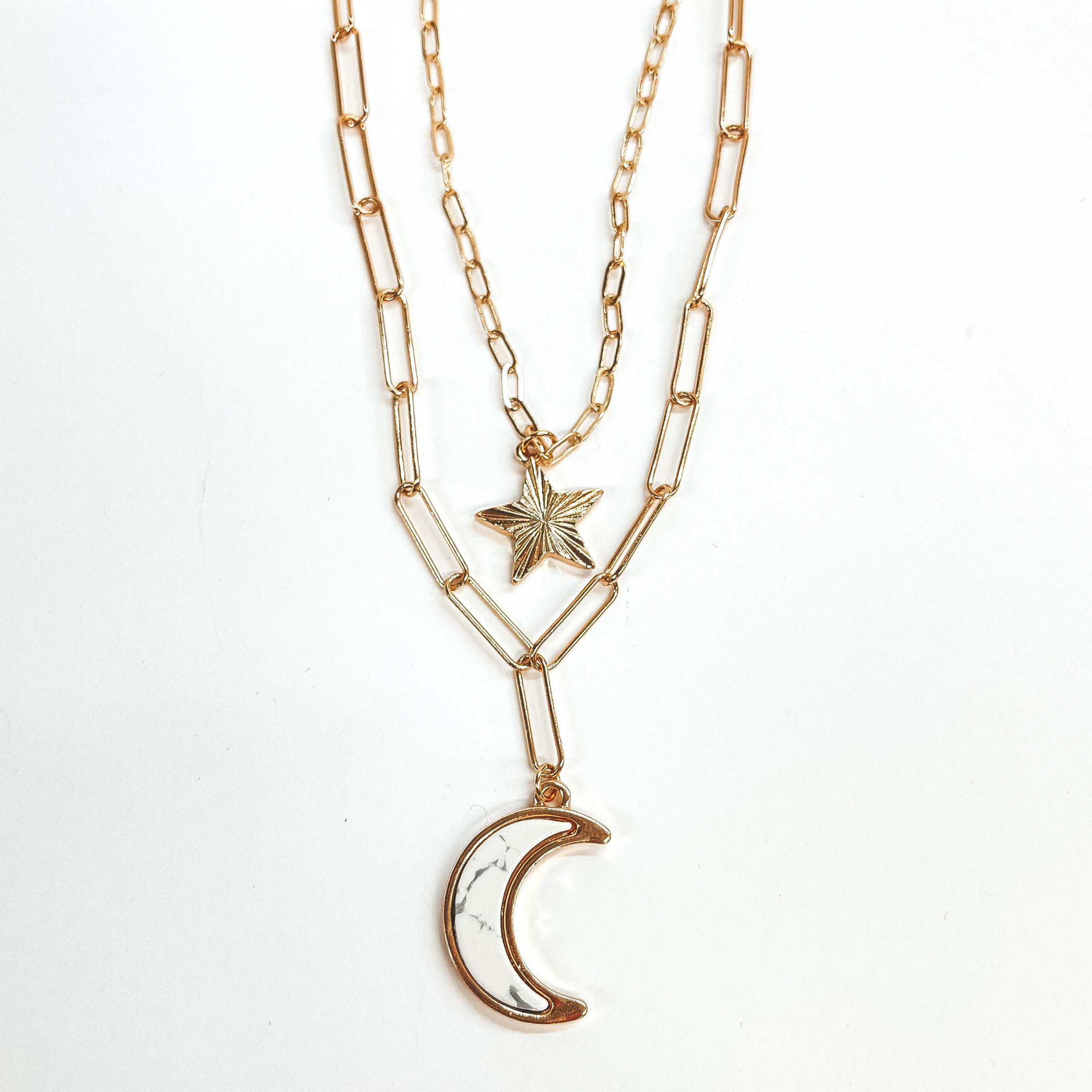 Double layered paperclip chain necklace in gold.  The shorter strand has a gold sunburst star and  the longer strand has a white stone pendant in a moon shape. This necklace is taken on a white background.