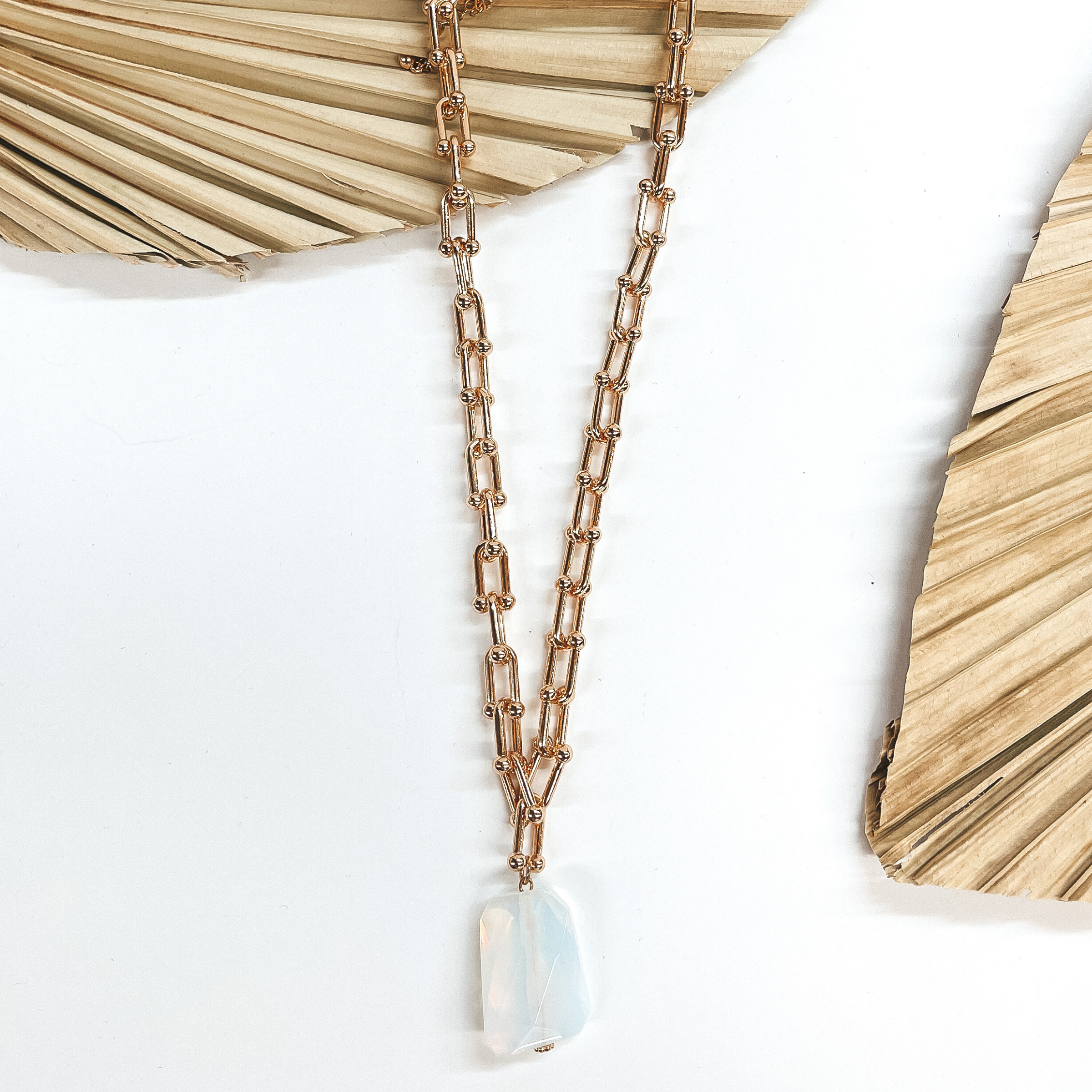 Gold thick link chain necklace with a big opal  iridescent stone pendant in the center. The necklace is taken laying on a dried up palm leaf and white background, with  another dried up palm leaf in the side as decor.