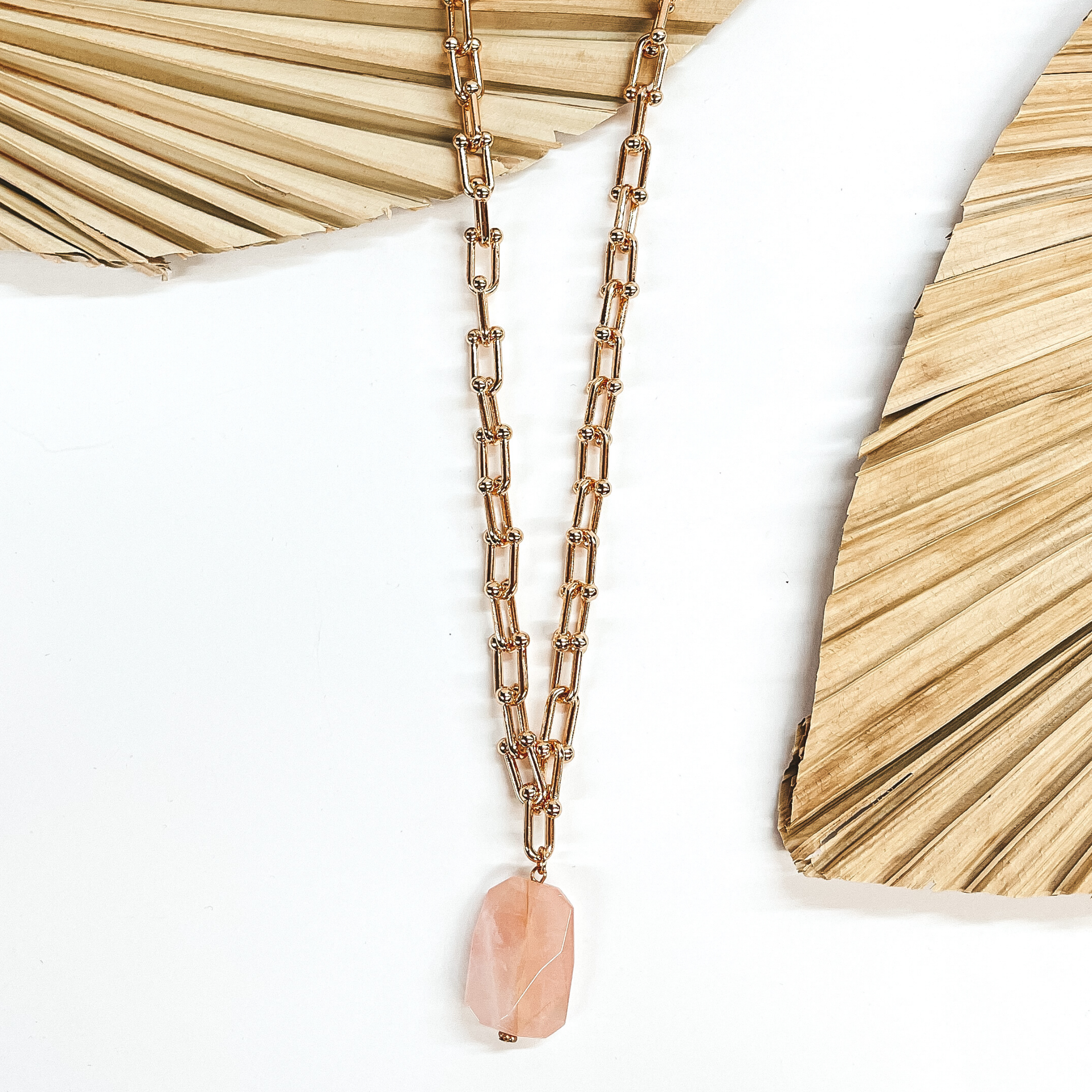 Gold thick link chain necklace with a big rose quartz pendant in the center. The necklace is taken laying on a dried up palm leaf and white background, with  another dried up palm leaf in the side as decor.