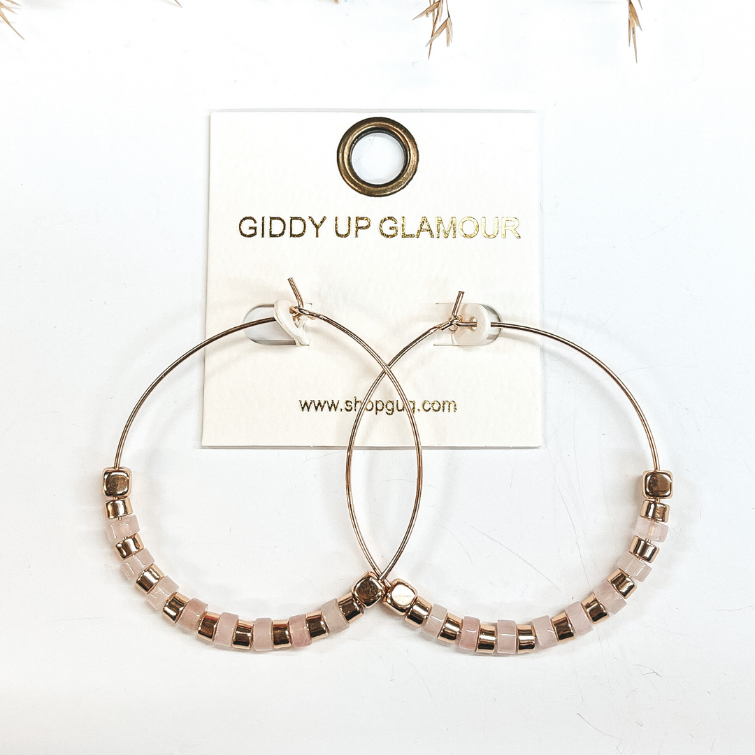 Gold hoops with gold and pale pink beads, on each end  there is a gold square bead. Taken on a white  background with a brown plant in the back as decor.