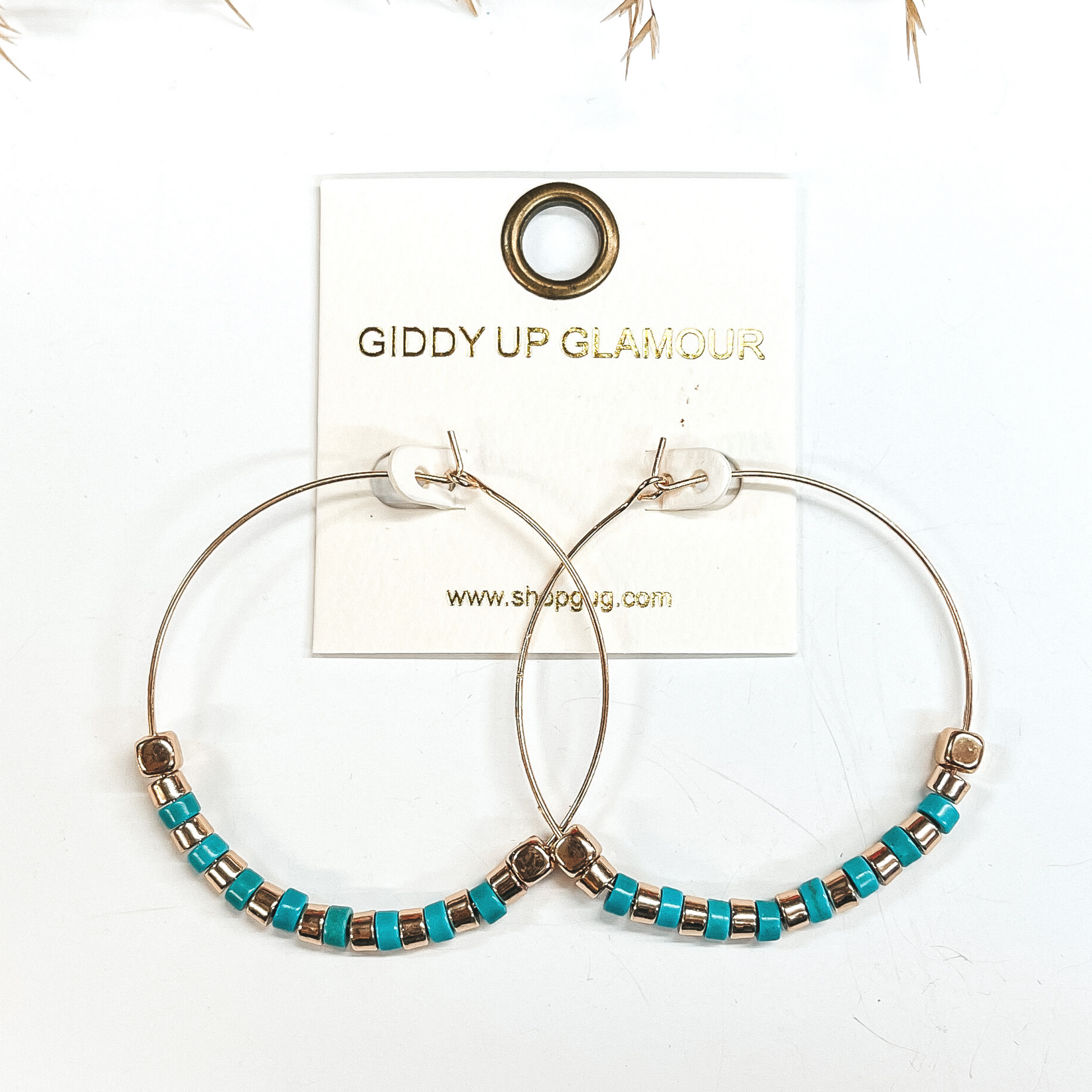 Gold hoops with gold and turquoise beads, on each end  there is a gold square bead. Taken on a white  background with a brown plant in the back as decor.