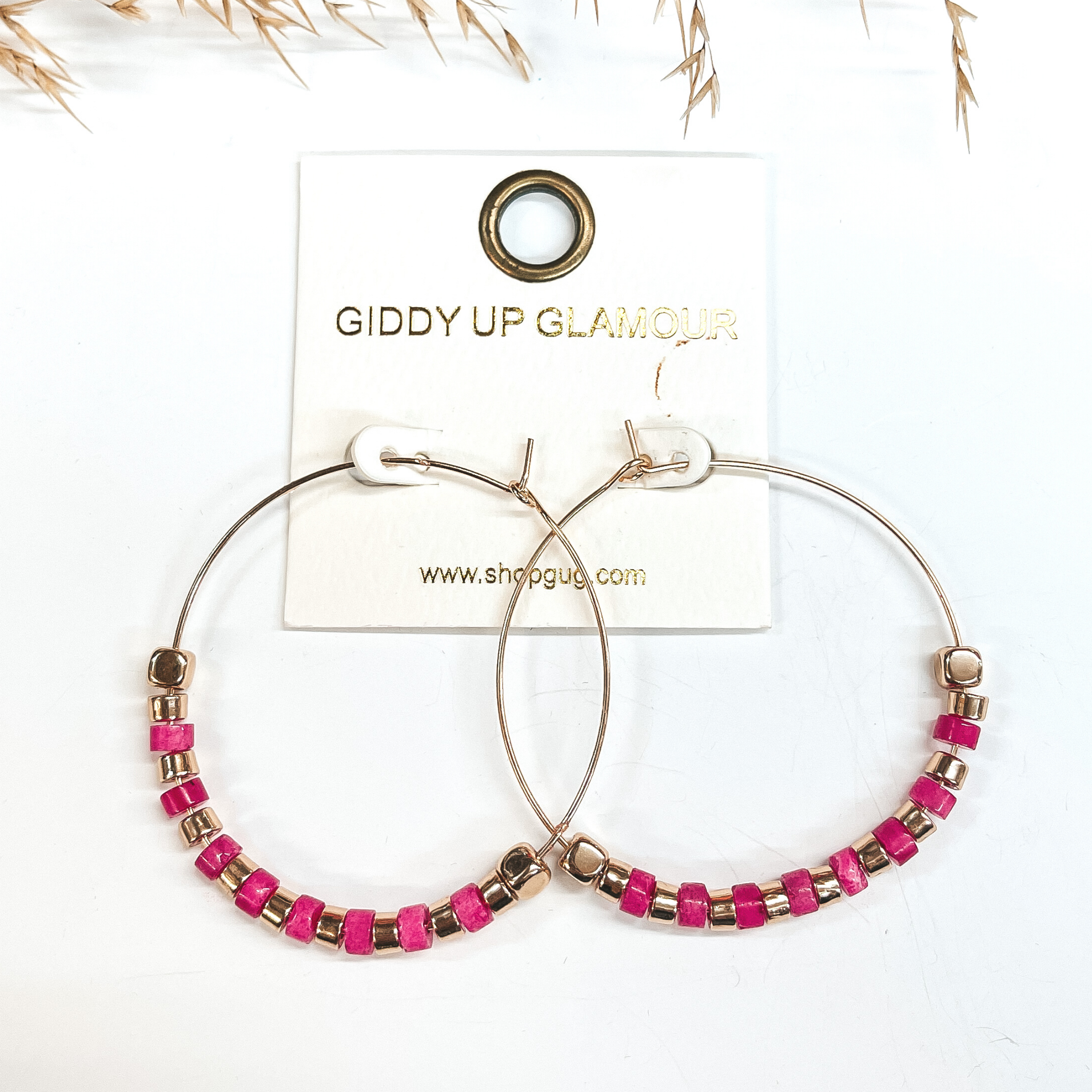 Gold hoops with gold and fuchsia beads, on each end  there is a gold square bead. Taken on a white  background with a brown plant in the back as decor.
