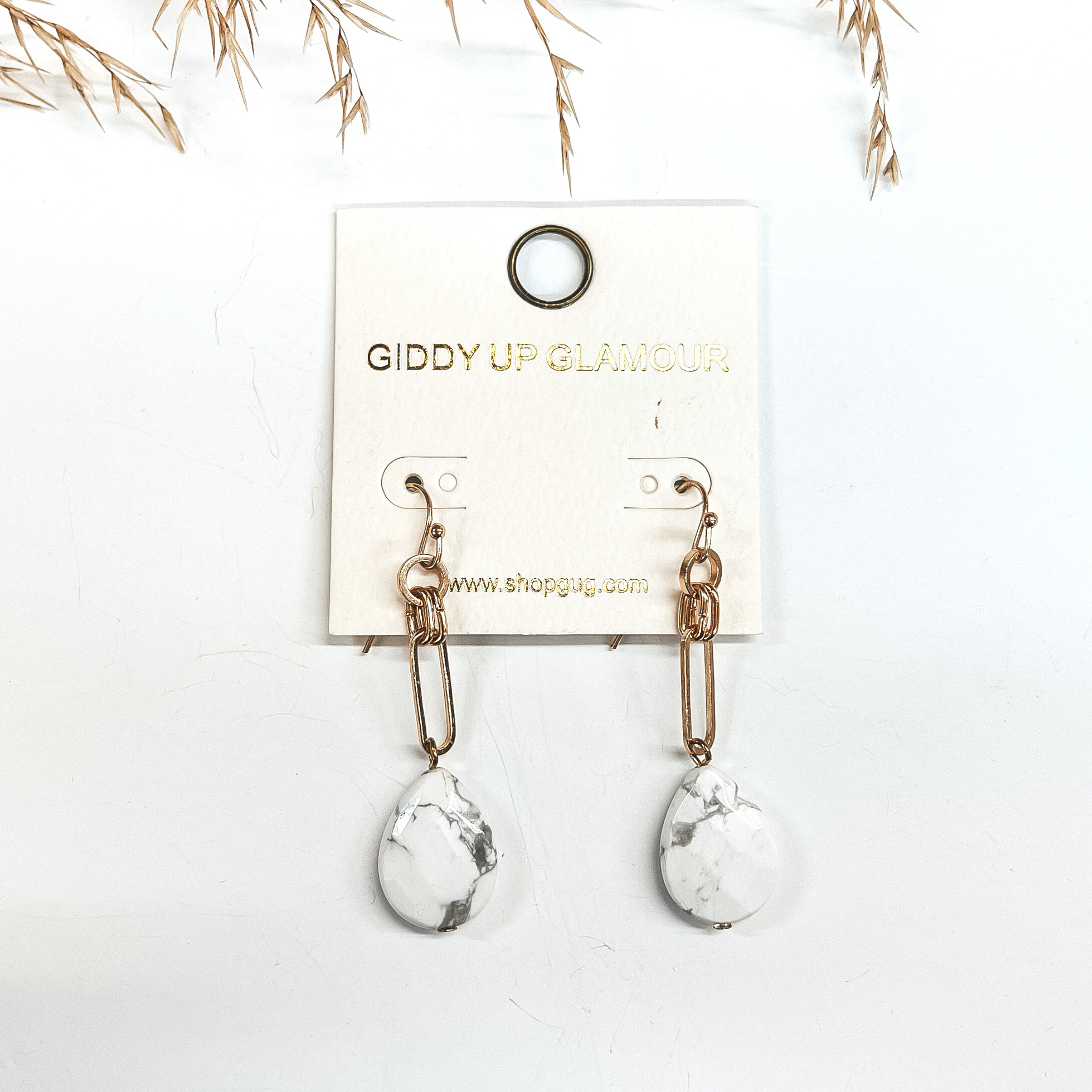 Gold chain earrings with a faux white stone drop. Taken on a white background with a brown plant in the  back as decor.