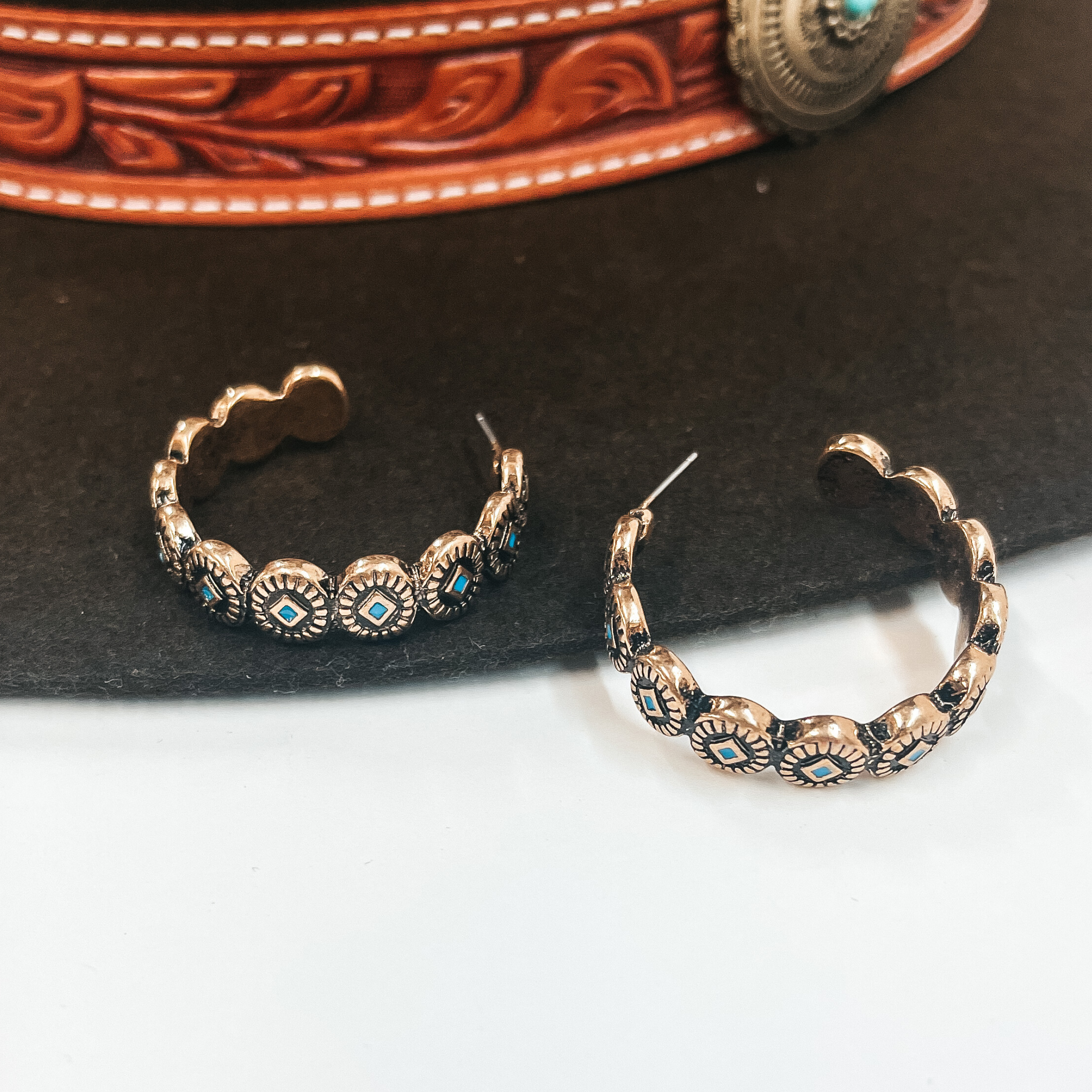 Gold semi hoop with post back backing, hoops are  engraved with western design and has a bit of  turquoise detailing as well. These earrings are taken  on a dark brown hat and white background. 